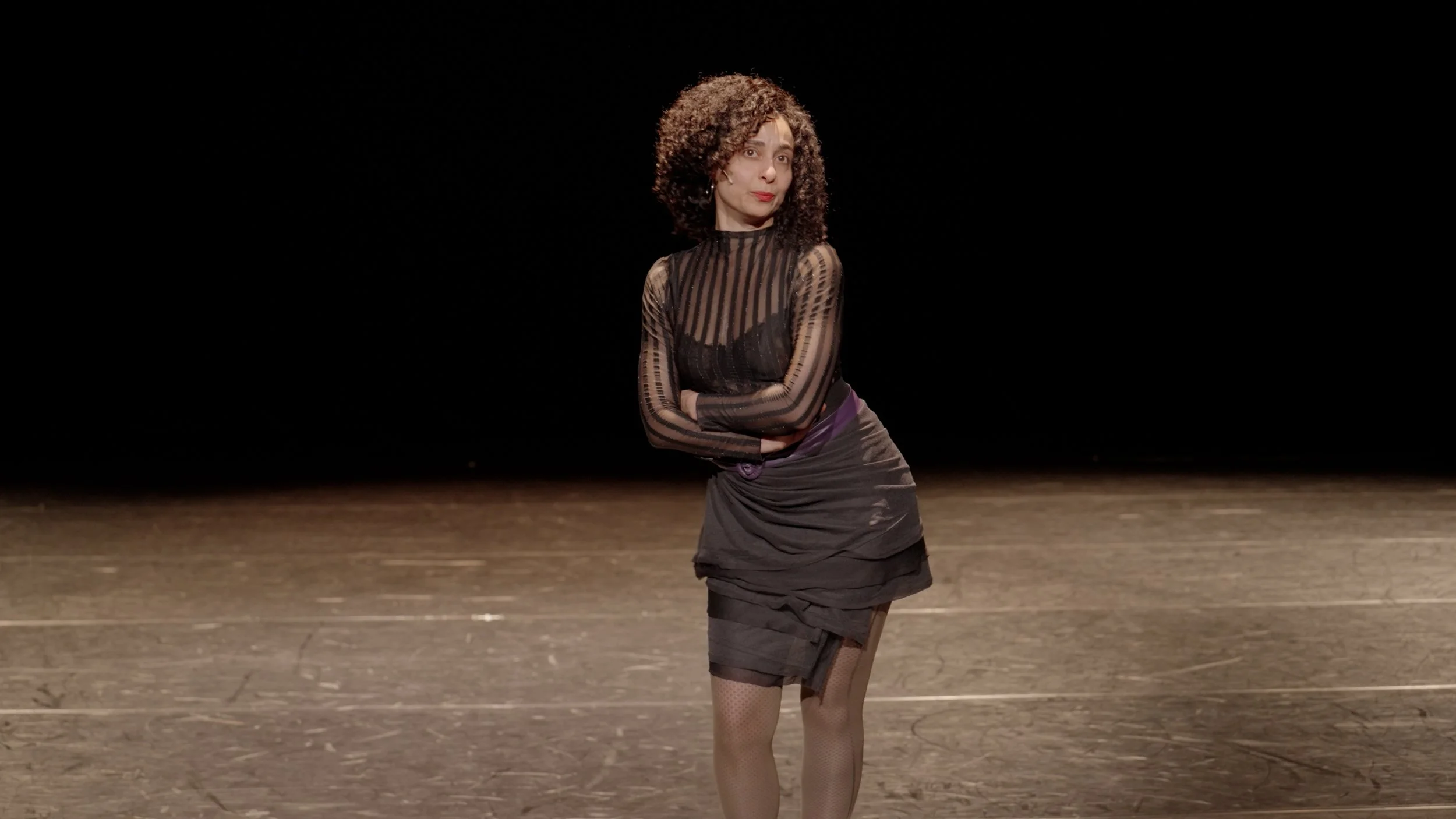 A woman with curly hair wearing a sheer striped black top, black skirt, and fishnet stockings stands on a dark, empty stage with arms crossed.