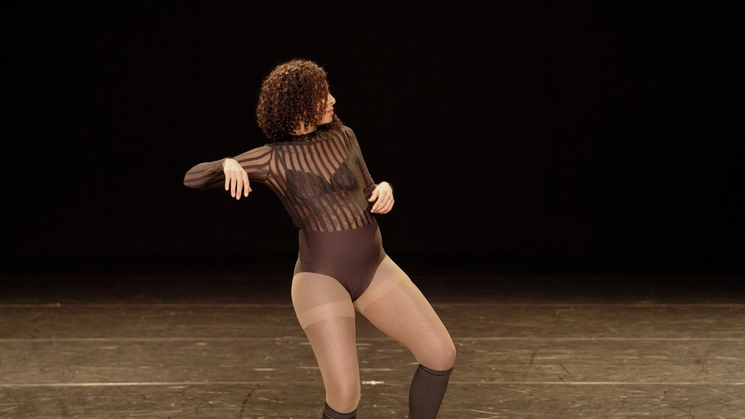 A woman with curly hair dancing on stage in a sheer, striped black top, black shorts, fishnet tights, and knee-high socks against a black background.