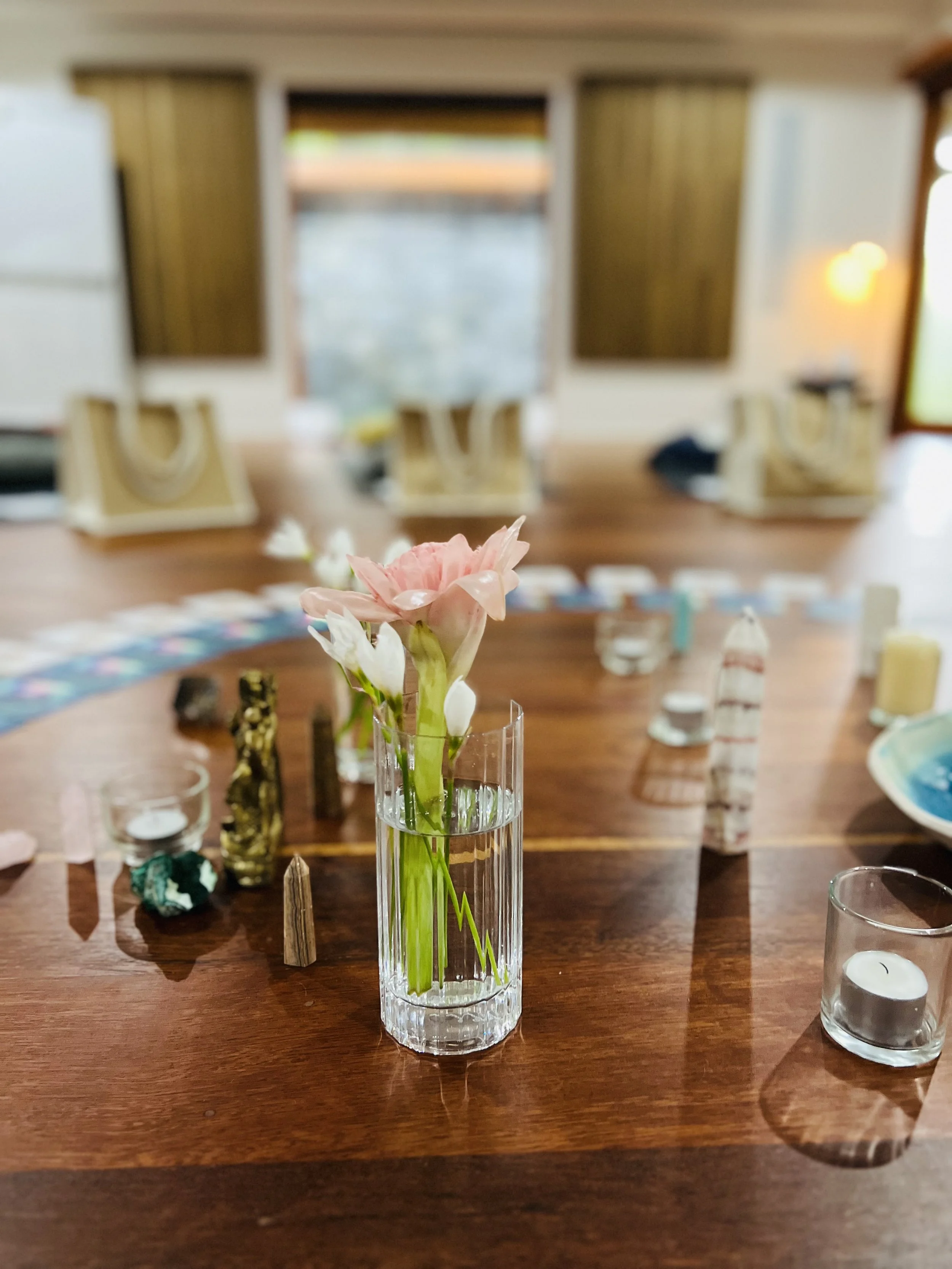 A pink flower in a tall clear glass vase on a wooden table, with various decorative objects and candles around it, and a blurred background of a room with windows and furniture.