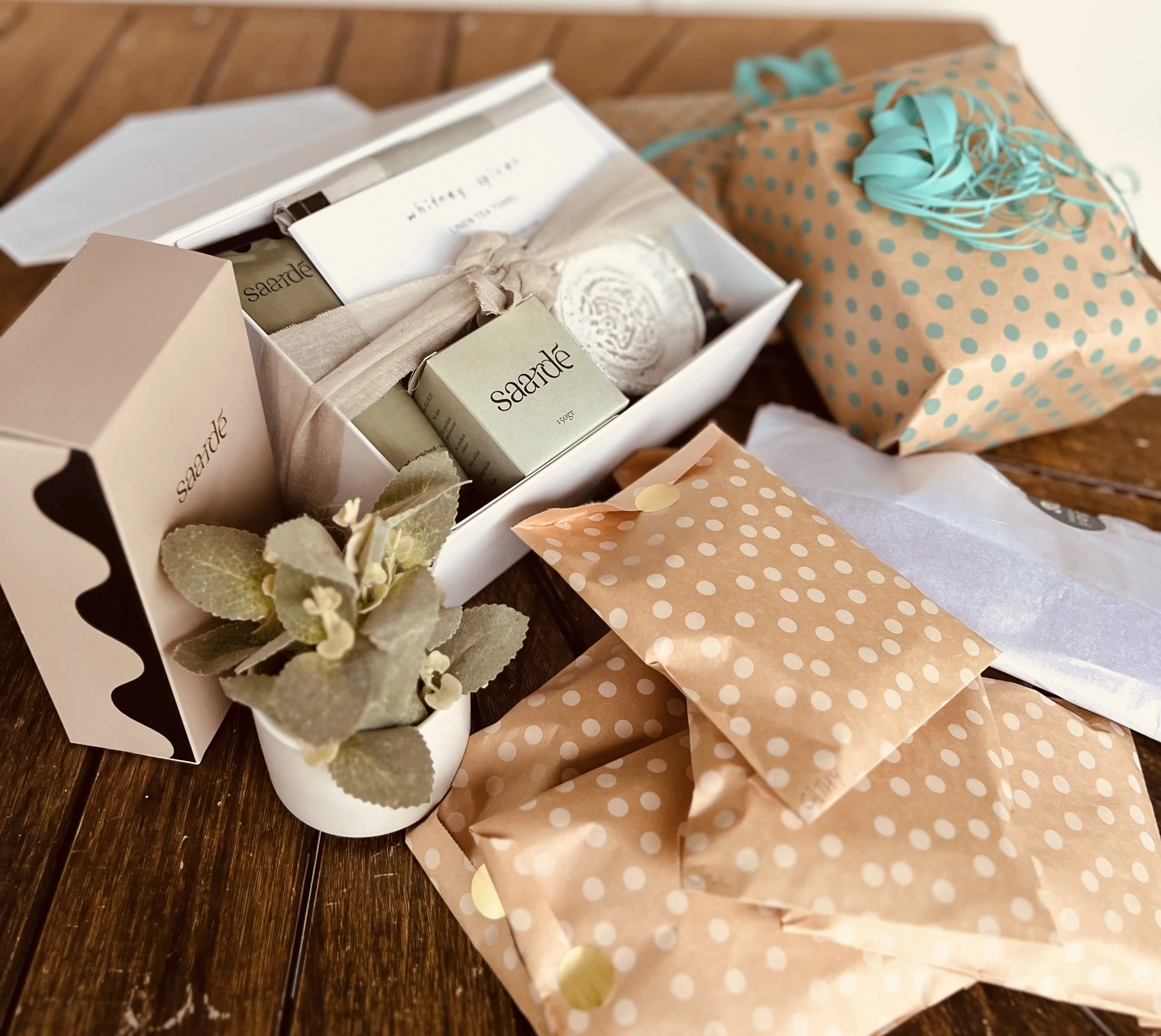 A gift set with soap, a box, and a plant in a small white pot, along with wrapped presents with pastel polka dot wrapping paper and ribbon, and tissue paper on a wooden surface.