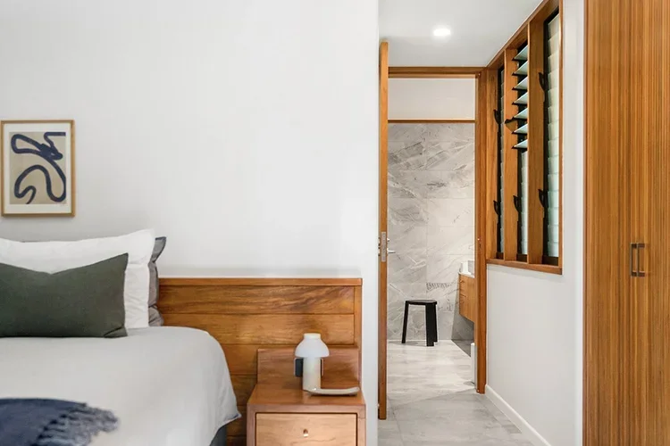 Modern hotel room with a large bed, blue blanket, white pillows, wooden headboard, gray armchair, nightstand with lamp, framed artwork on the white wall, and wood flooring.