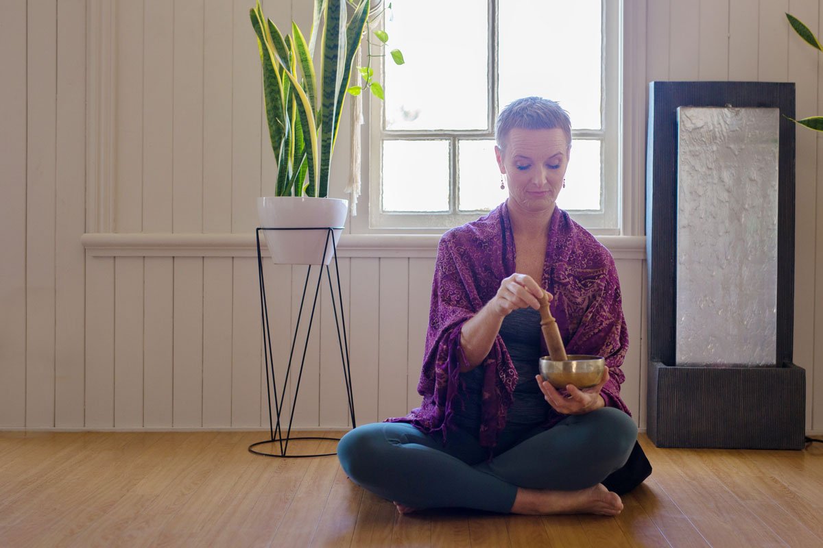 A woman sitting cross-legged on wooden floor, meditating and using a singing bowl with a mallet, in a bright room with window, potted plant, and mirror.