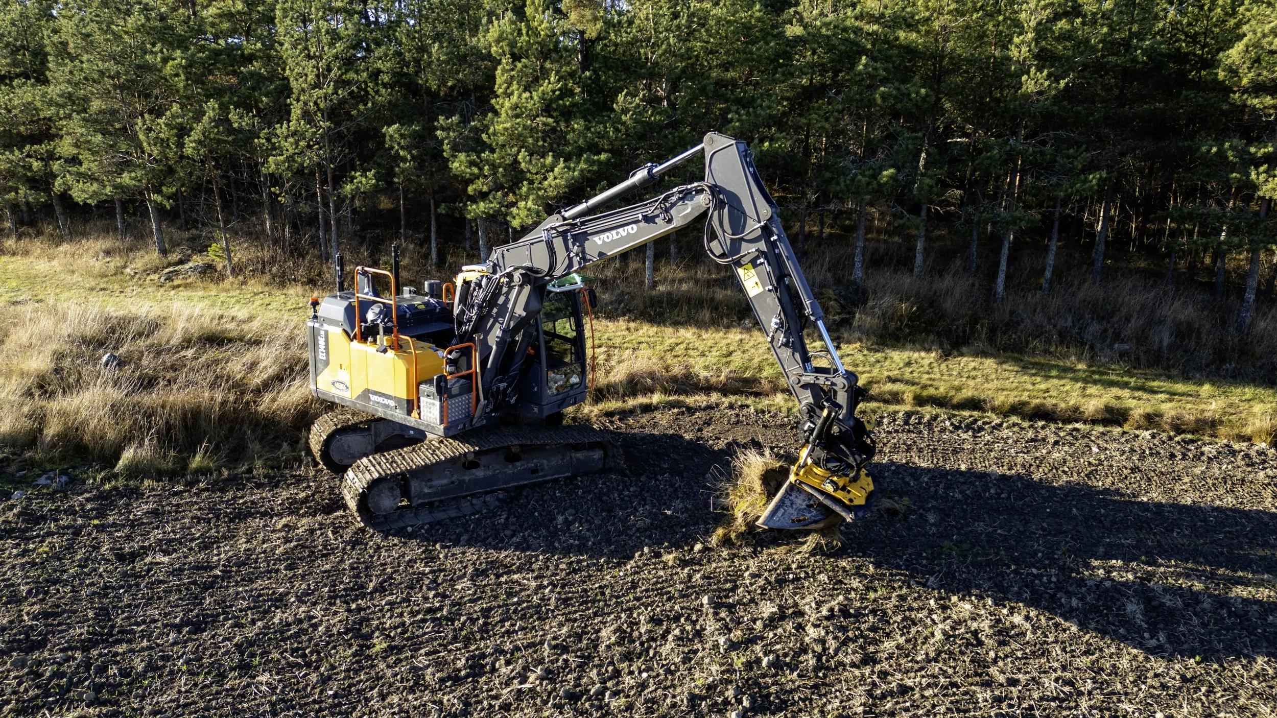 En gul och svart grävmaskin som gräver i jorden i ett öppet fält med träd i bakgrunden.