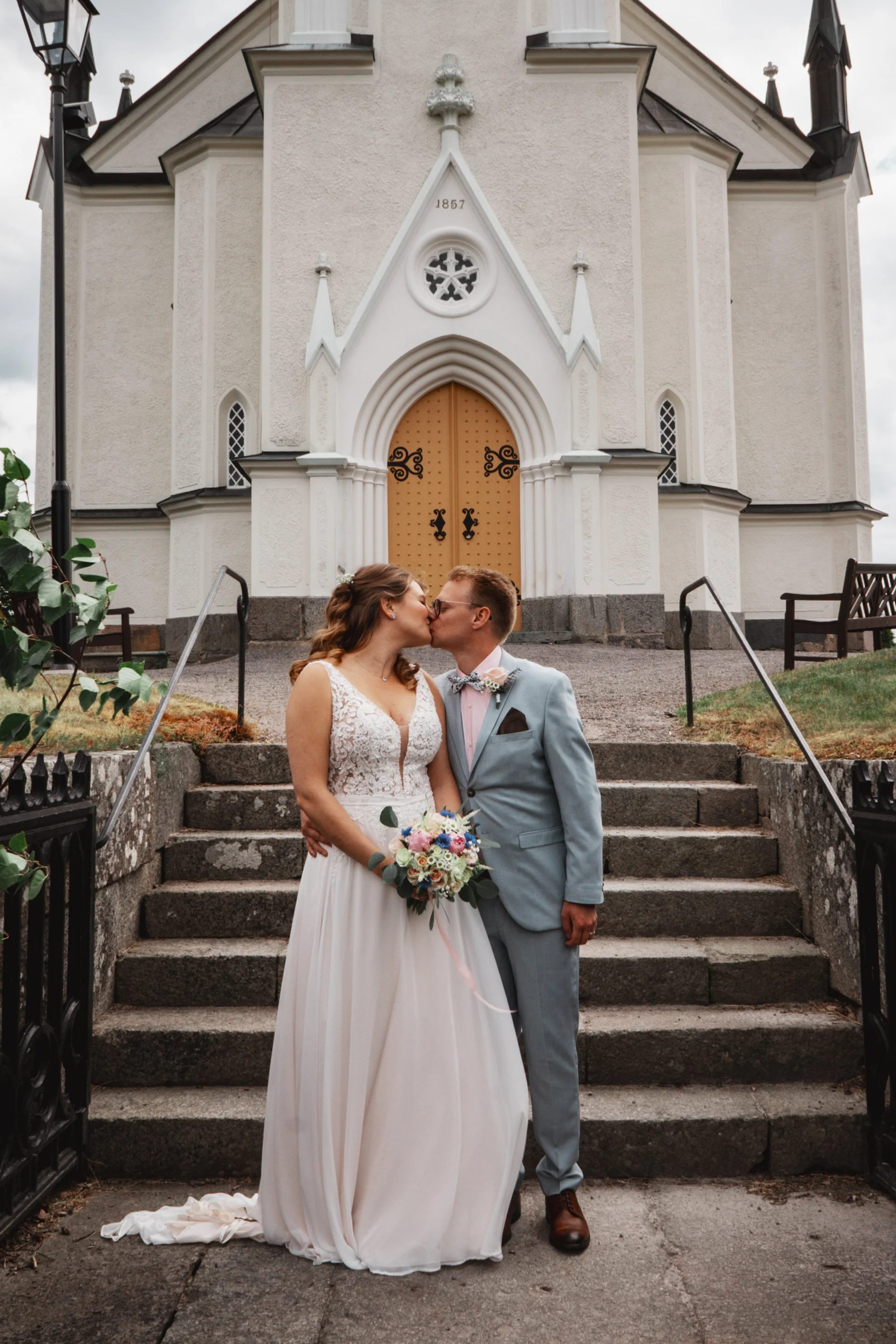 Nygift par kysser framför en vit kyrka, kvinnan håller en bukett blommor, mannen har på sig en ljusblå kostym, kyrkans utgång är trappa med stenar.
