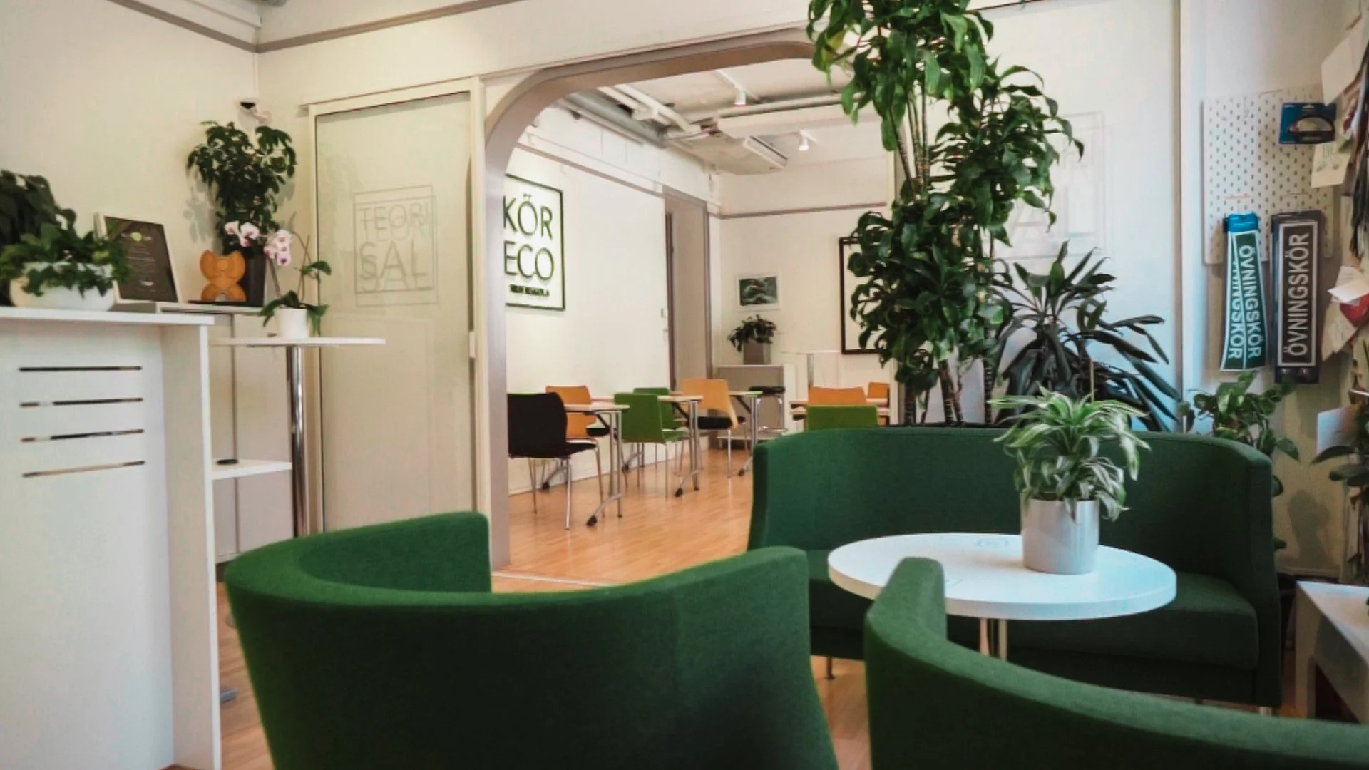 Interiör av ett grönt och vitt mötes- eller caféområde med gröna växter, soffor, bord och stolar i olika färger, samt skyltar och bilder på väggarna.