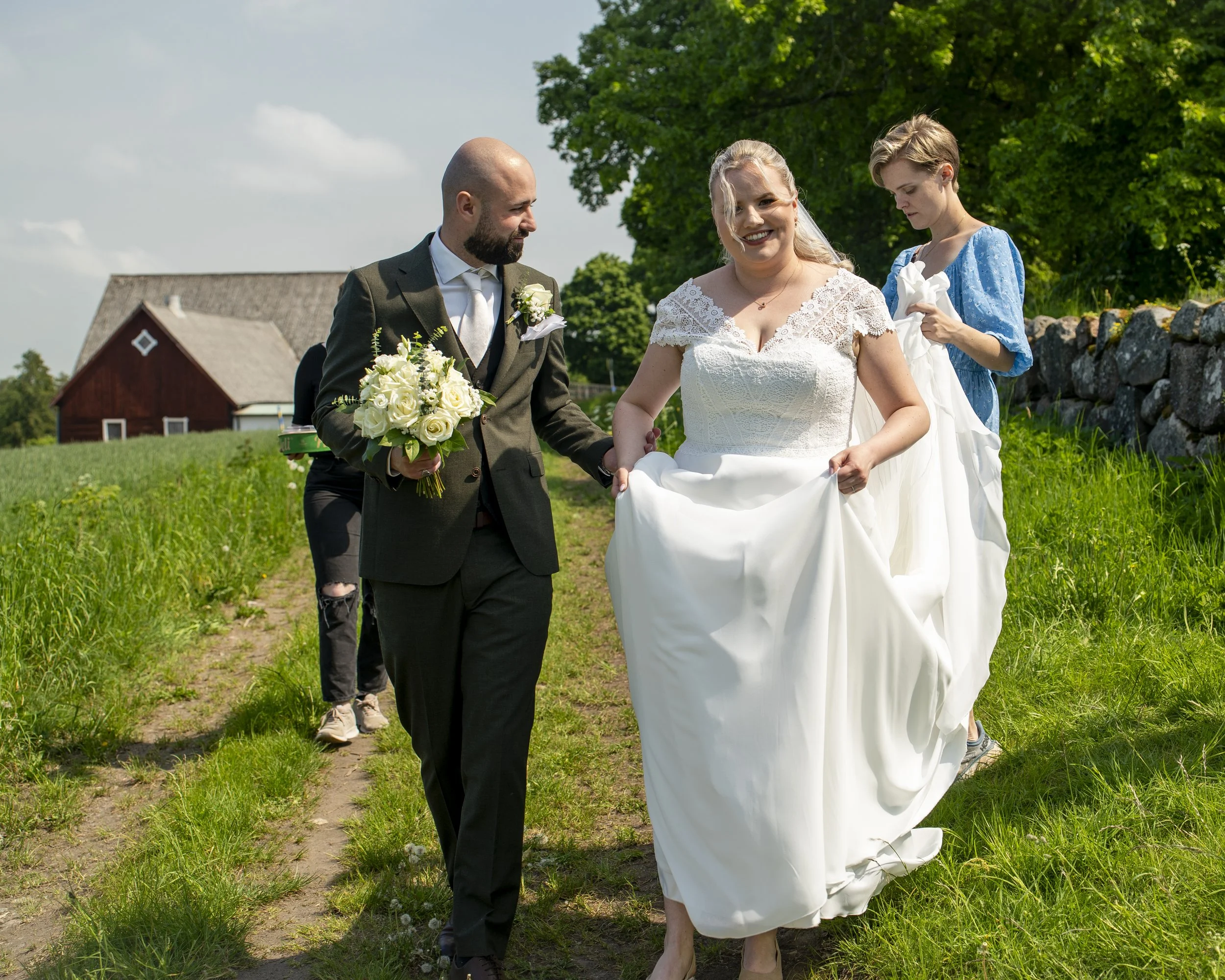 Brudparet går ute på landet med en kvinna som hjälper med bröllopsklänningen, på en grön grusväg, när solen skiner.