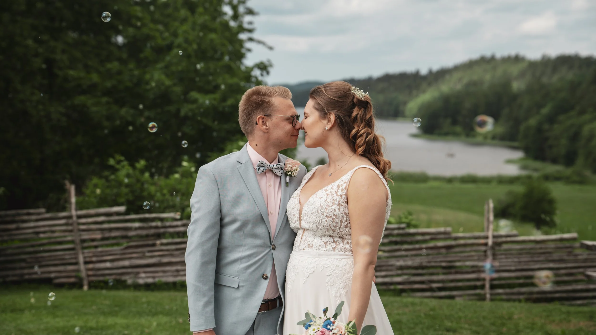 Nygifta paret står nära varandra utomhus, med en sjö och skog i bakgrunden, och är på väg att kyssa varandra på bröllopsdag.