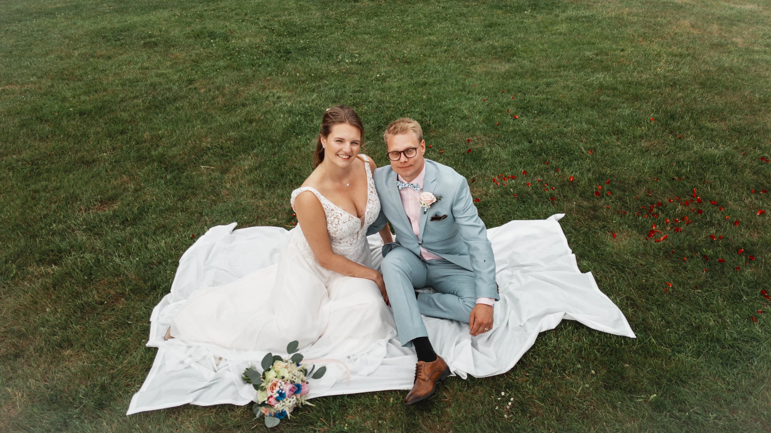 Nybrollopsparet sitter på en vit duk på gräset, omringad av röda blomblad, med en bukett blommor framför dem.