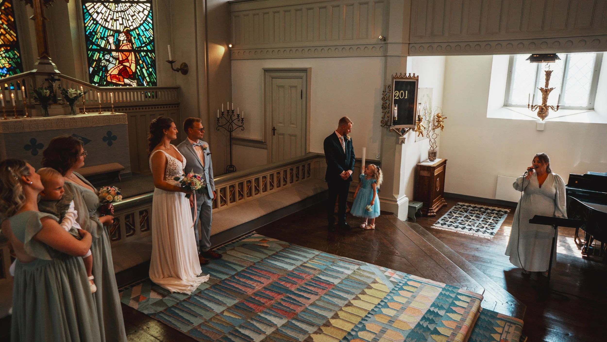 Vid ett altare i en kyrka håller ett brudpar, med omgivande gäster, ett bröllopslöfte inför prästen. Brun trägolv, färgglad matta, fönster med ljusinsläpp, och ett stort kupolfönster med kors på.