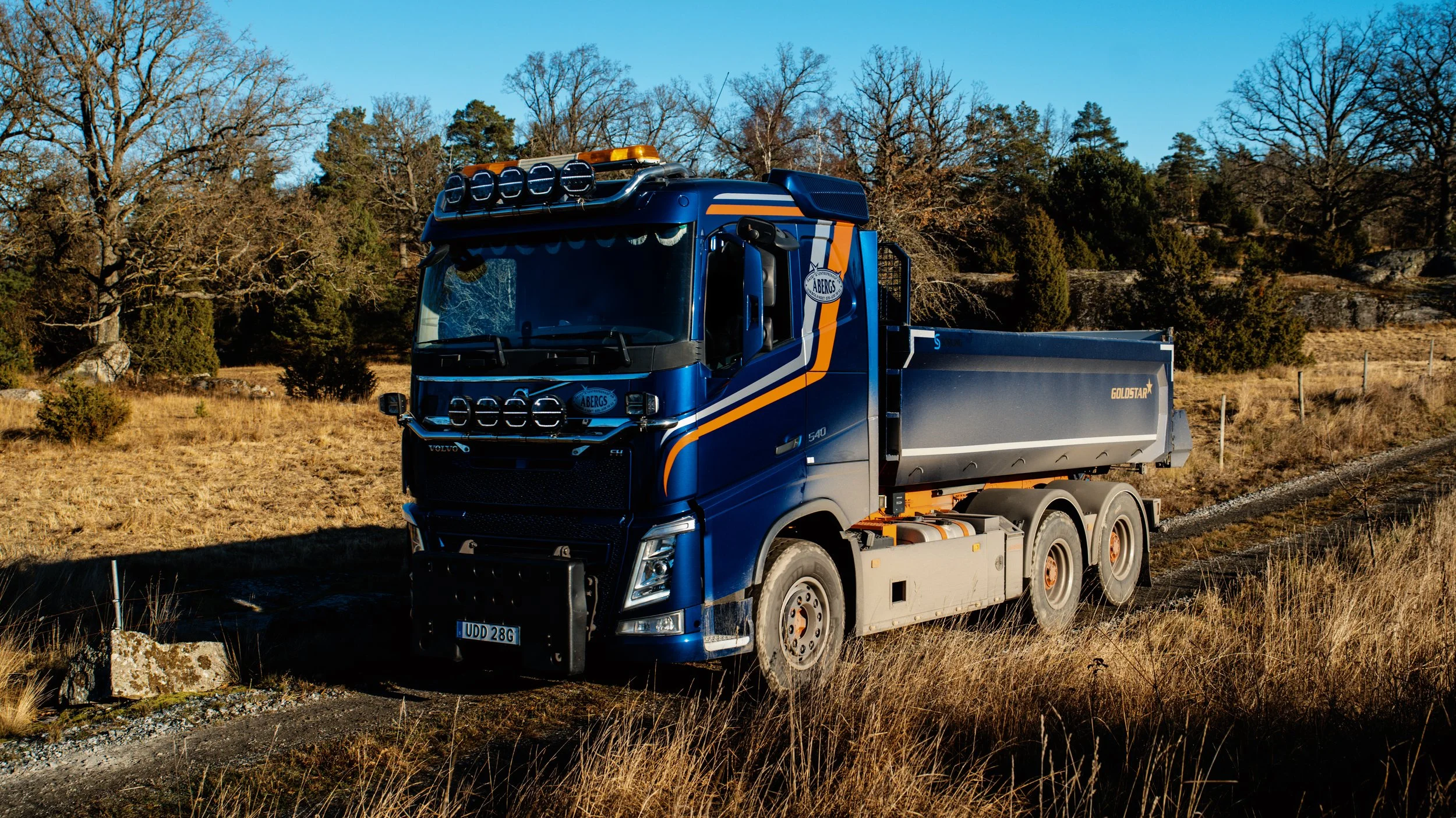 En blå lastbil kör på en jordig väg i ett öppet landskap med träd i bakgrunden, under en klar himmel.