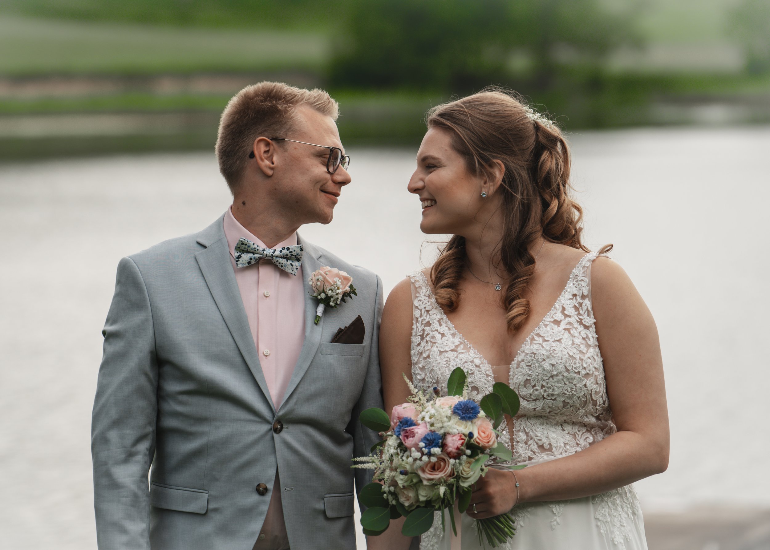 En brud och en brudgum i bröllopskläder som står nära varandra vid vattnet, ler mot varandra, utomhus.