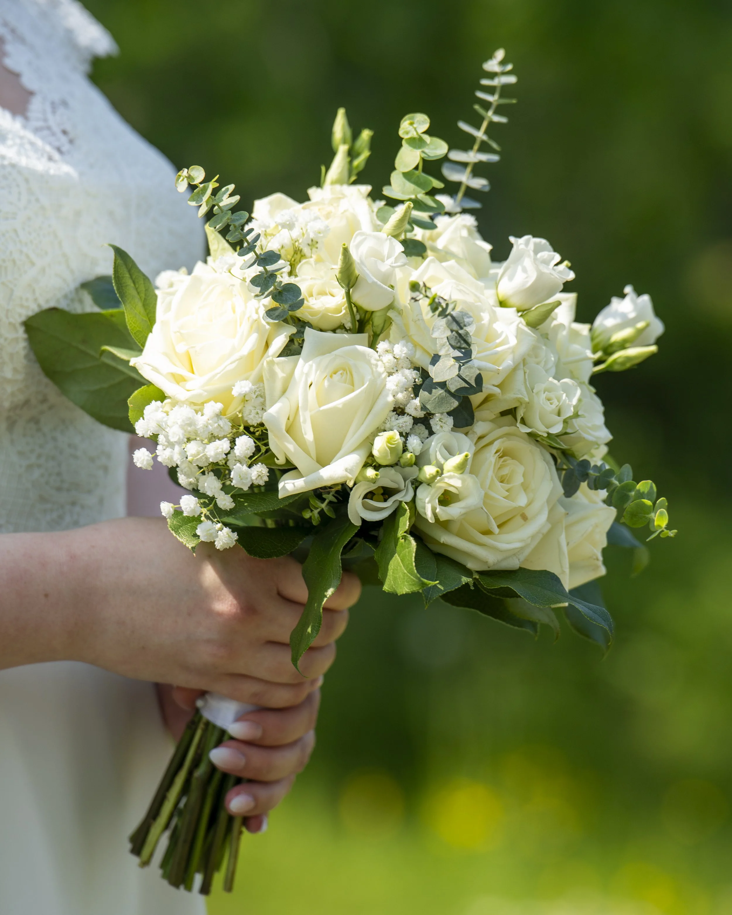 En person håller en bukett av vita blommor, inklusive rosor och eucalyptus, mot en grön bakgrund.