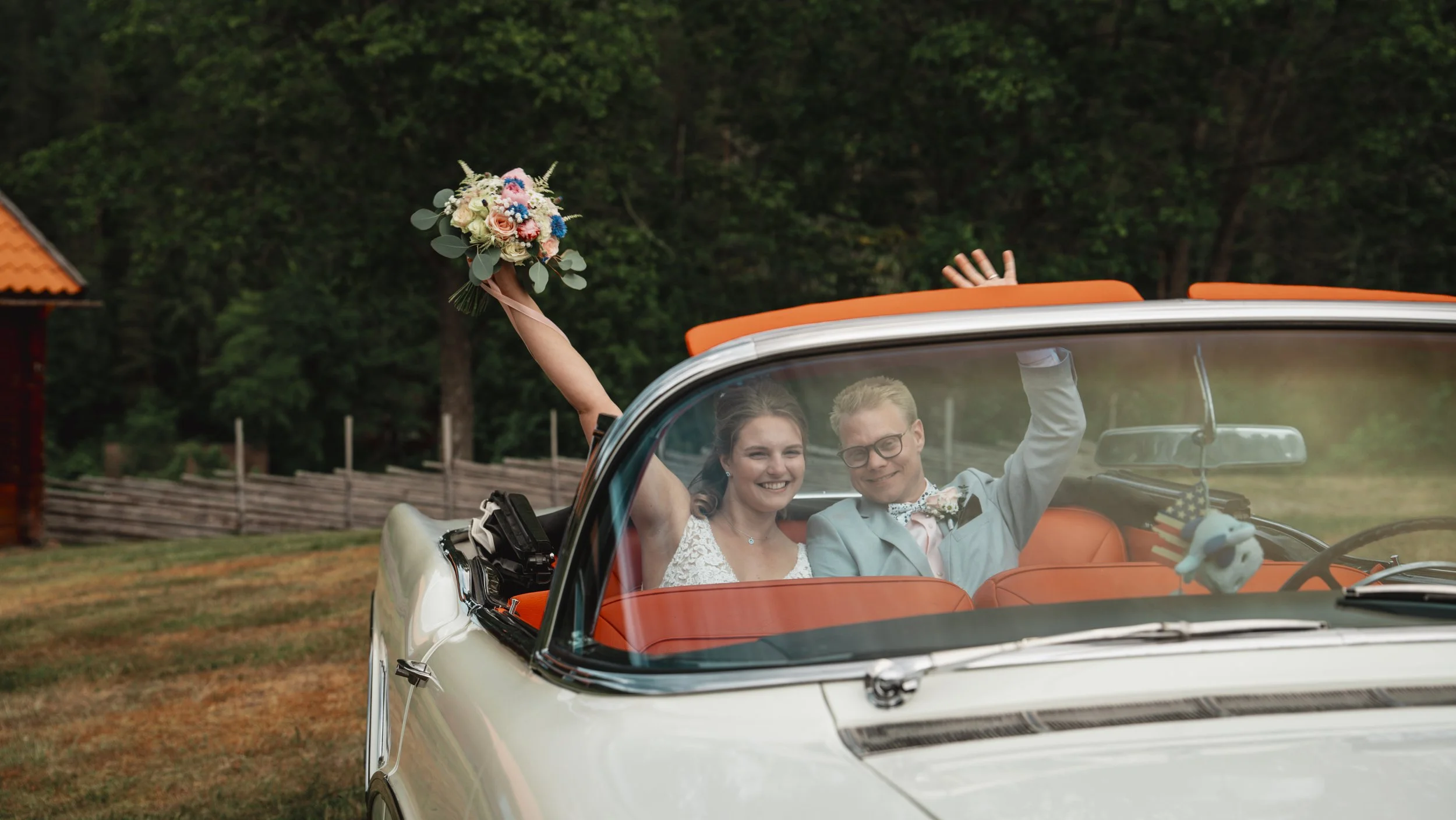 En lycklig brud och brudgum åker i en vit bil med orange inredning på landet, båda ler och vinkar, bruden håller en bukett i handen. Bakom dem syns en skog och en liten röd stuga.