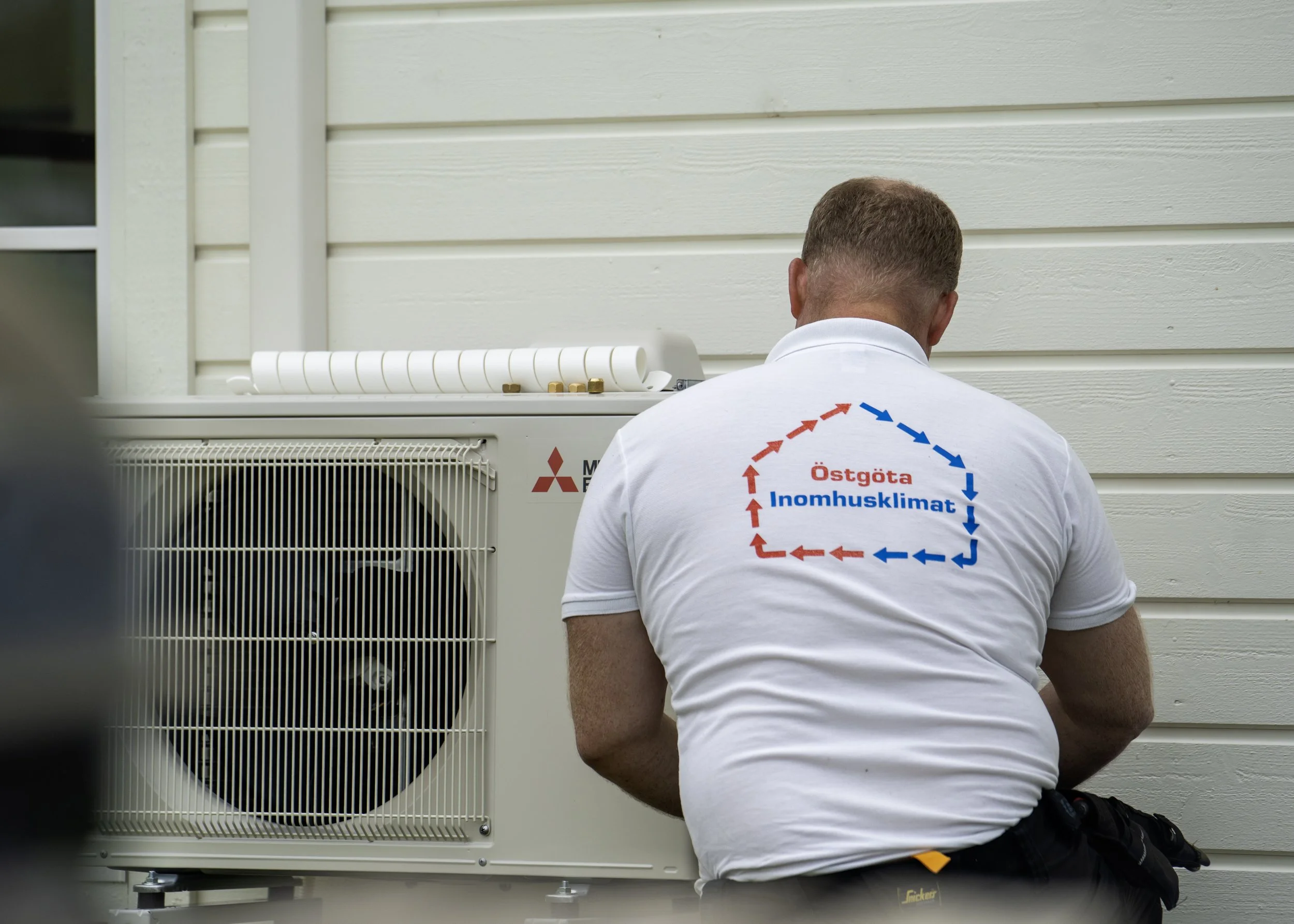 En man sitter på huk framför en utomhusenhet för luftkonditionering eller värmepump, sett bakifrån, med text på ryggen som säger "Östgöta Inomhusklimat".