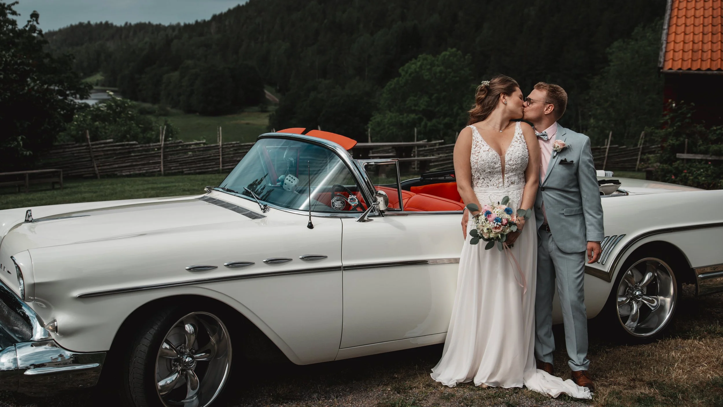 Nygifta par kysser framför en vit klassisk bil med röd inredning utomhus, omgivna av gröna träd och landskap.