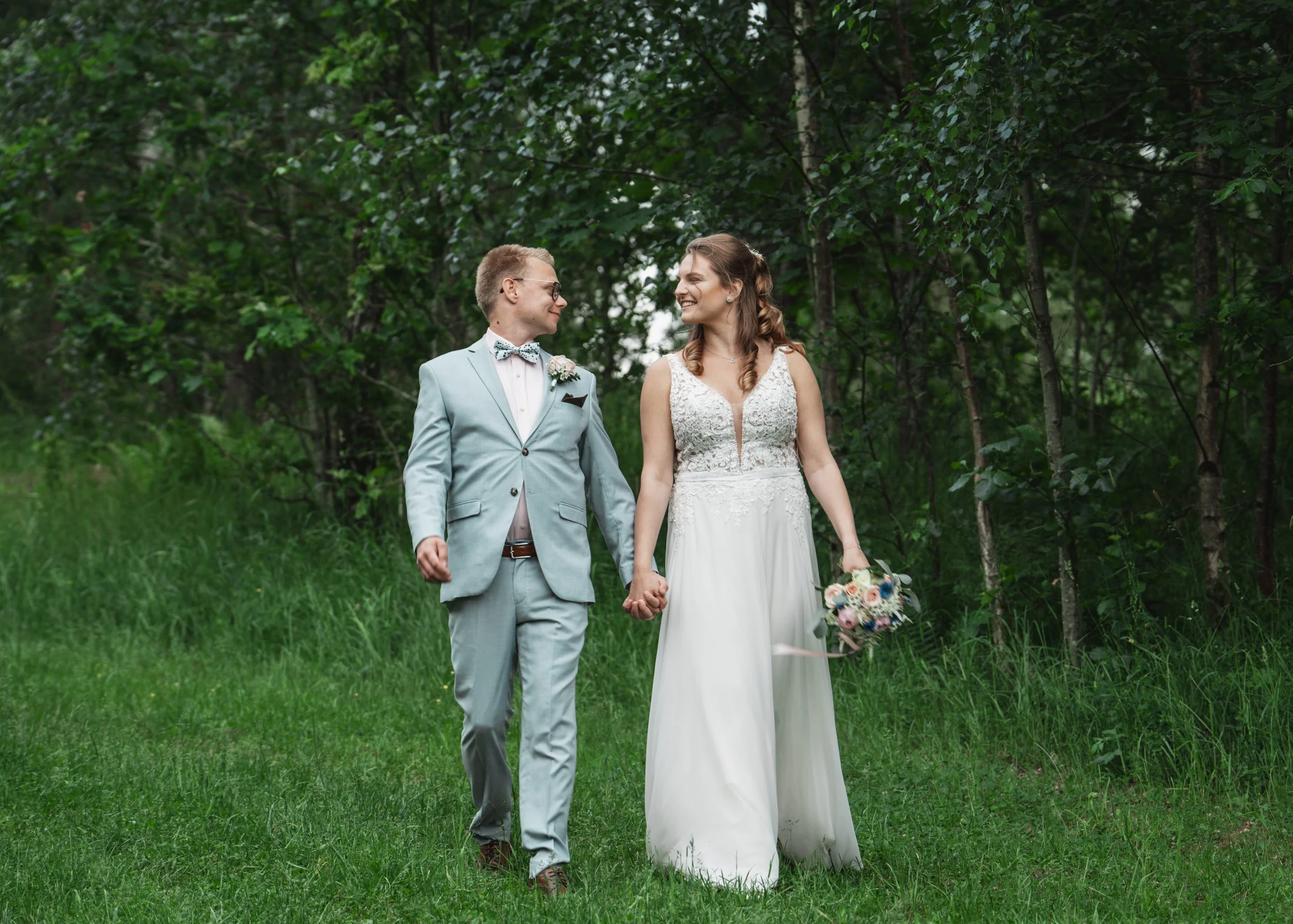 En nygift man och kvinna håller varandra i handen och går genom en grön skog, båda ler och tittar på varandra, kvinna bär en vit brudklänning och man ett ljusblått kostym, han har ett blombroder och hon håller en bukett blommor.