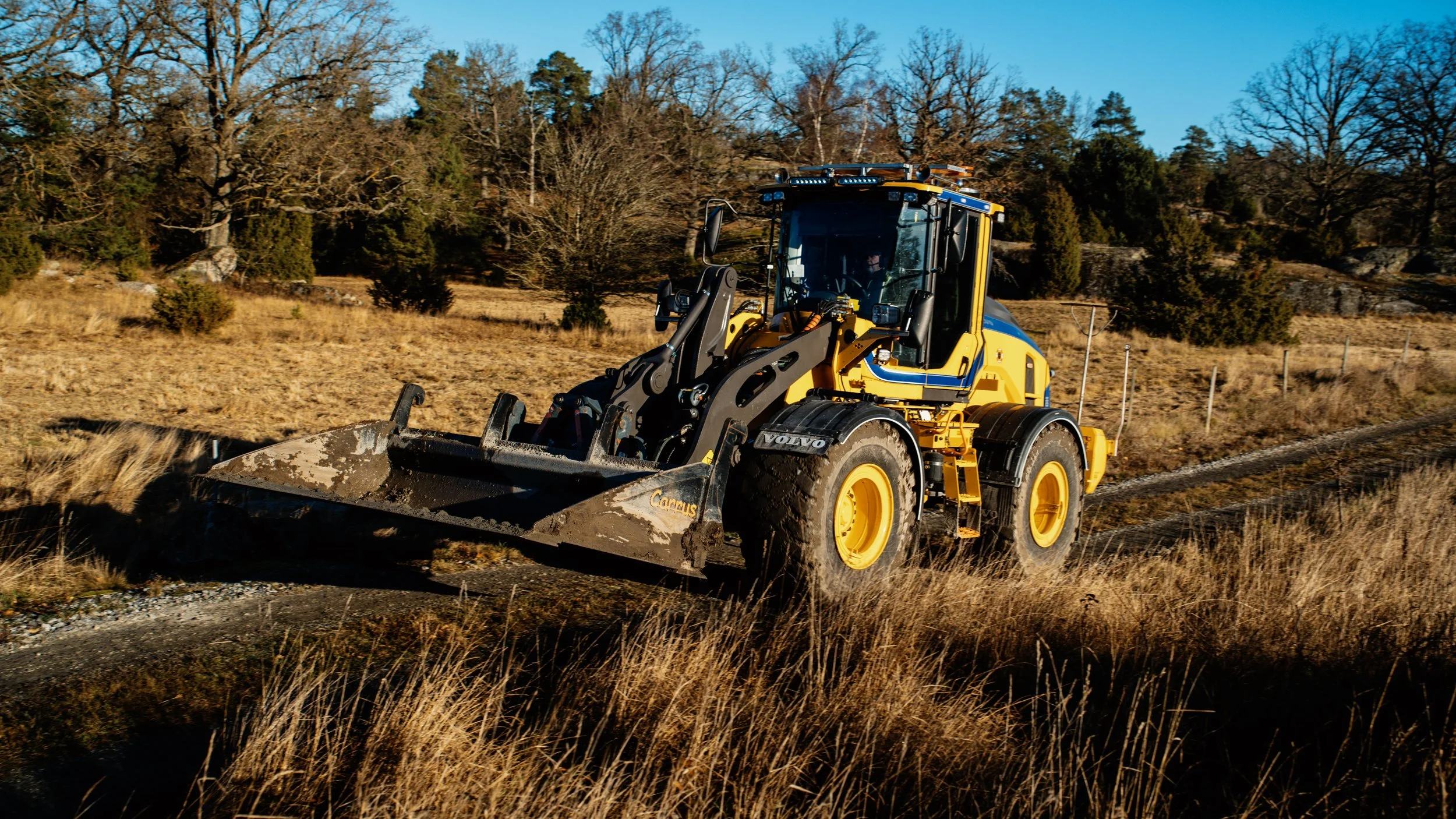 En gul och blå grävmaskin arbetar på en lantlig väg, omgiven av torra gräs och träd i bakgrunden.