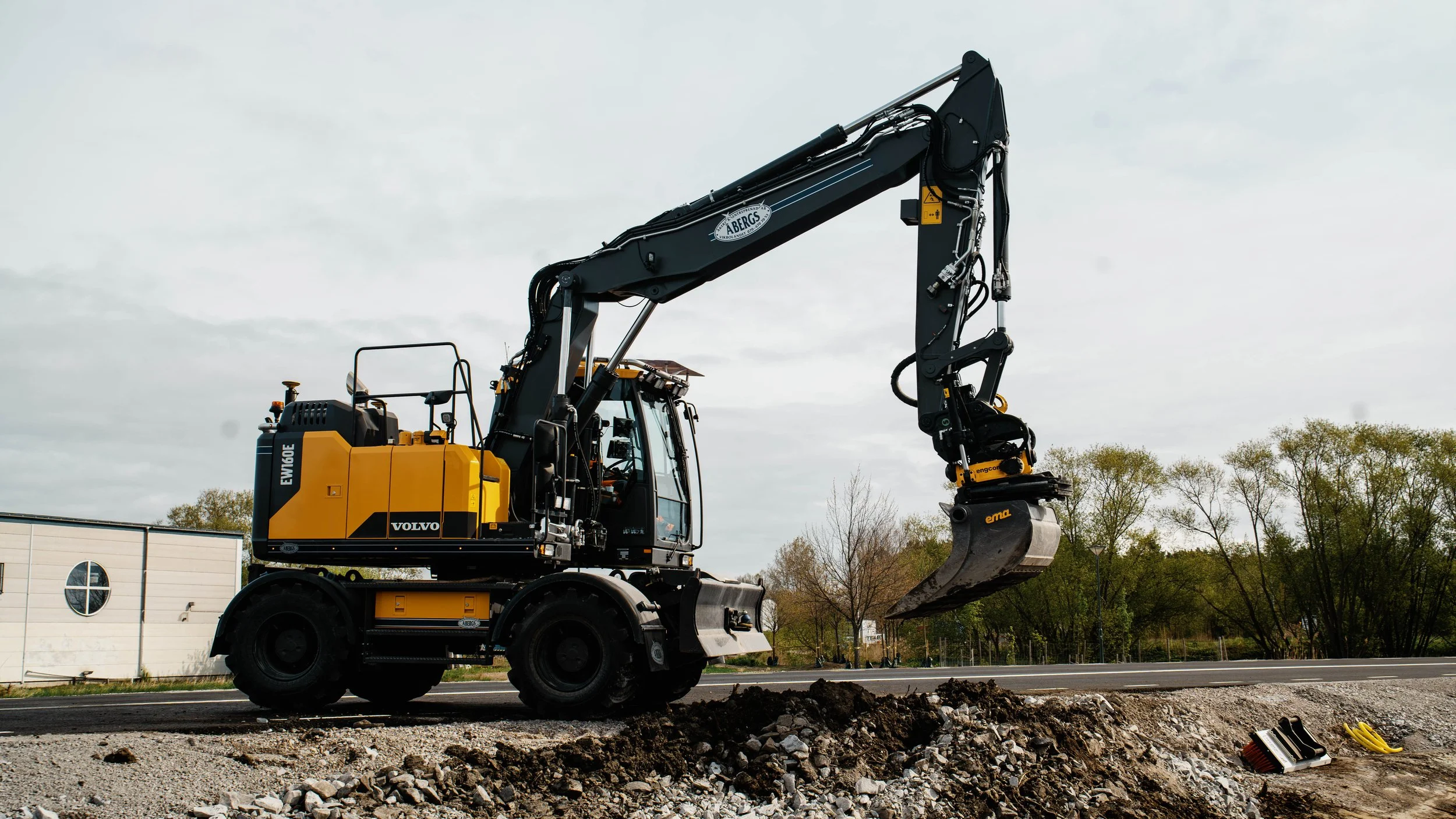 En gul och svart Volvo-grävmaskin som gräver vid en grusväg, med träd och en vit byggnad i bakgrunden.