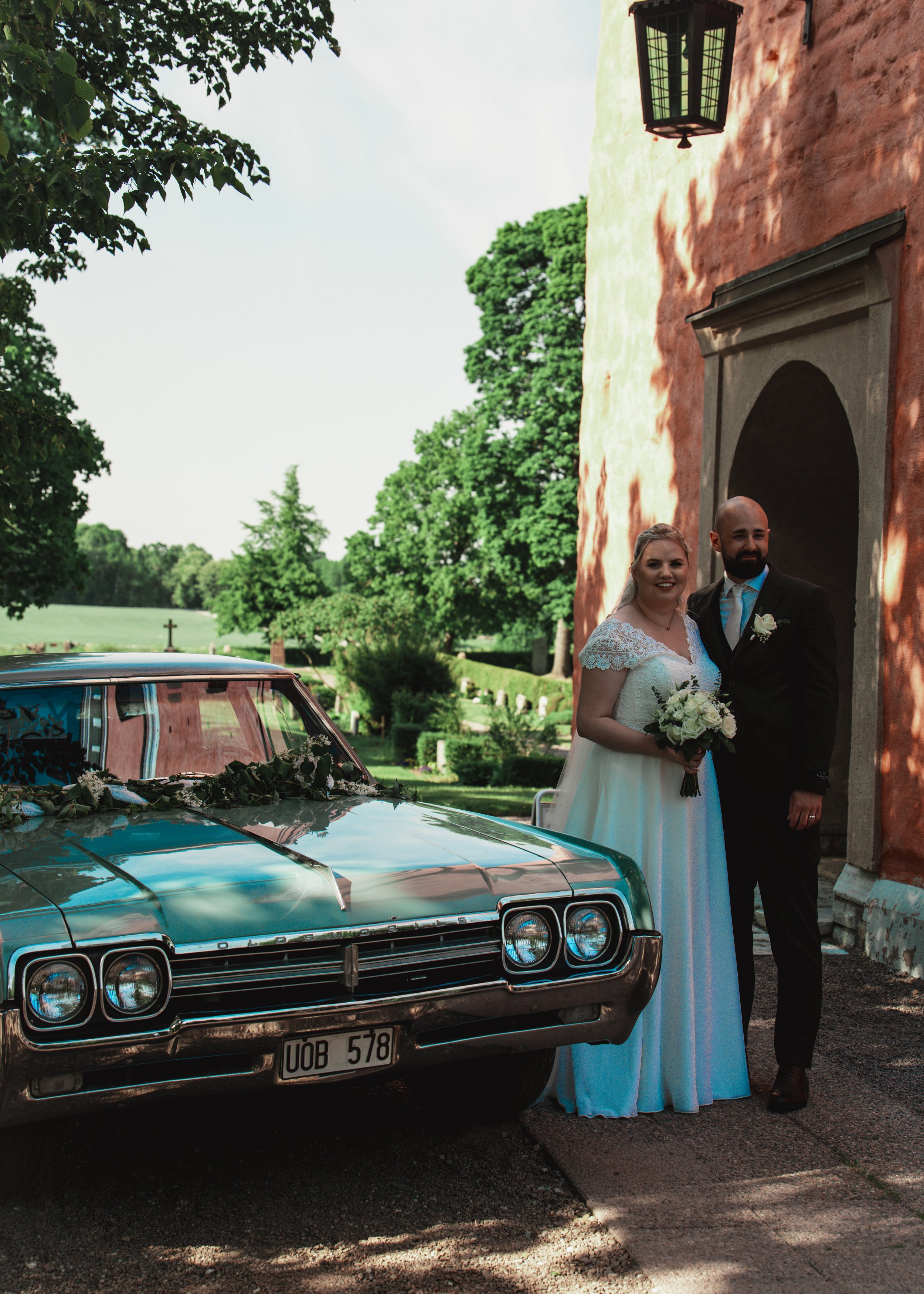 En brud och en brudgum står utanför en gammal byggnad med orange väggar, tillsammans med en klassisk bil dekorerad för bröllop. Bruden bär en vit klänning och håller ett blomsterarrangemang, medan brudgummen är klädd i svart kostym.