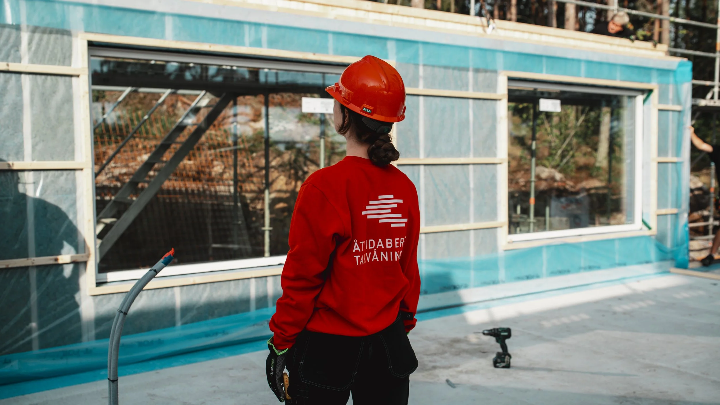 En kvinna i rött arbetslinne och orange hjälm står framför en byggnad under uppbyggnad. Det finns verktyg på golvet och byggmaterial i bakgrunden.