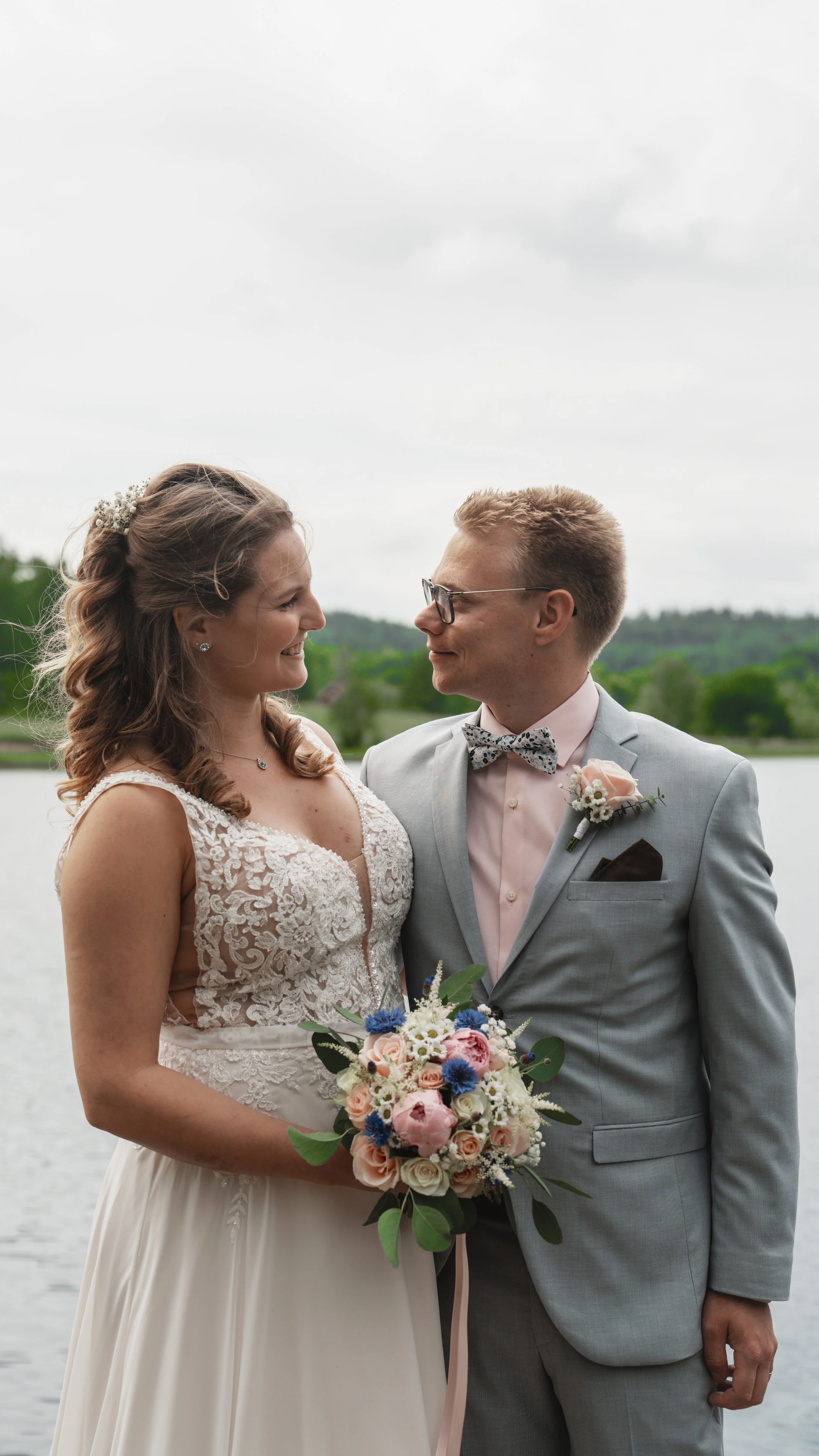 En brud och brudgummi står nära varandra utomhus vid vattnet, blickar in i varandras ögon, bruden håller i en bukett med rosa, vita och blå blommor.