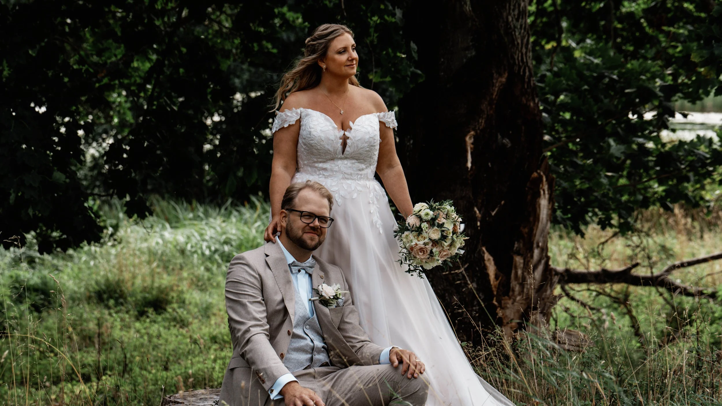 En brud i vit klänning håller en bukett och står bredvid en man i ljusgrå kostym i en naturlig, grön miljö.