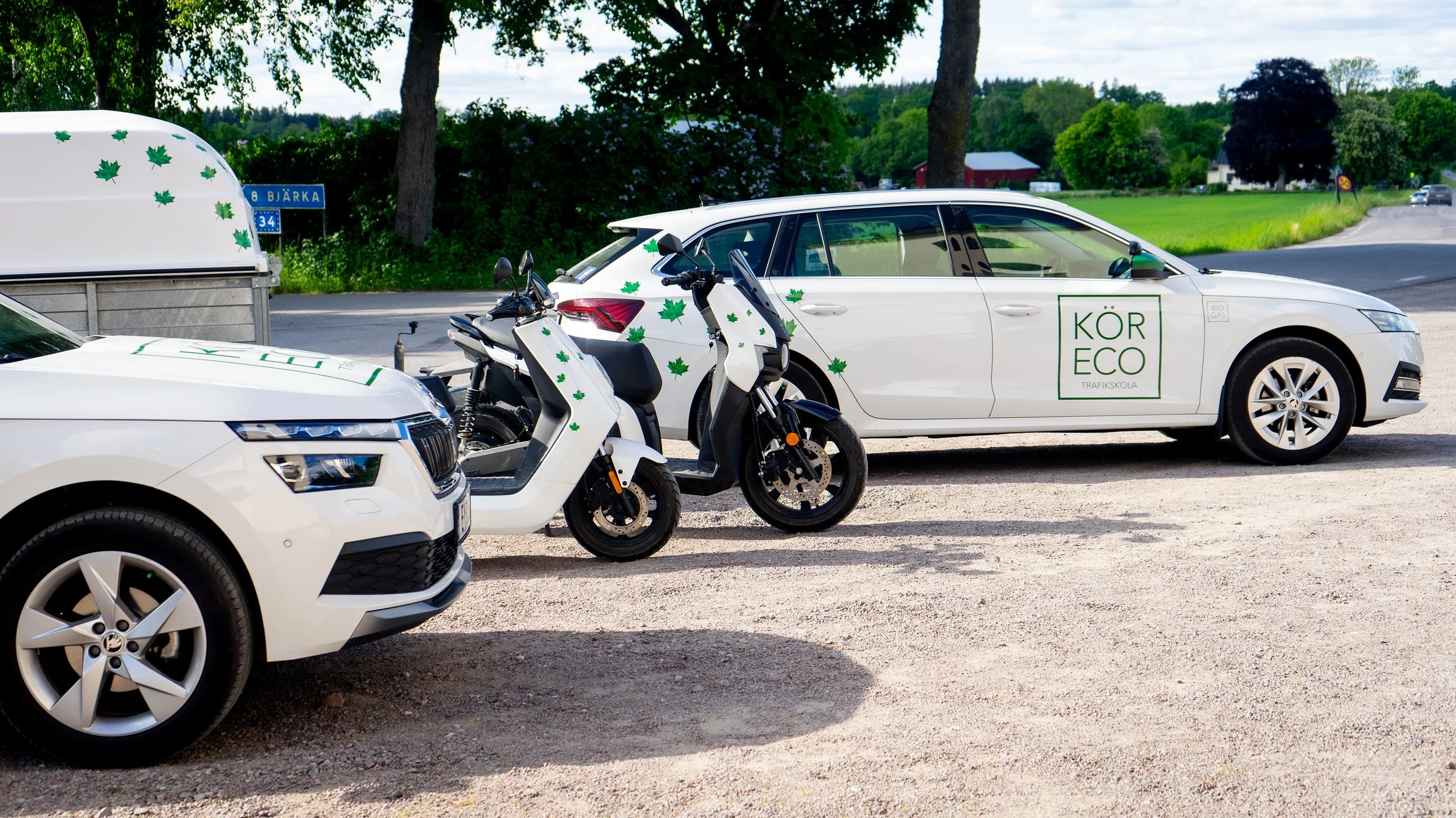 Tre vita fordon parkerade, en bil, en motorcykel och en scooter, alla dekorerade med gröna löv. En av bilarna har en etikett med 'KÖR ECO TRAFIKSKOLA'.