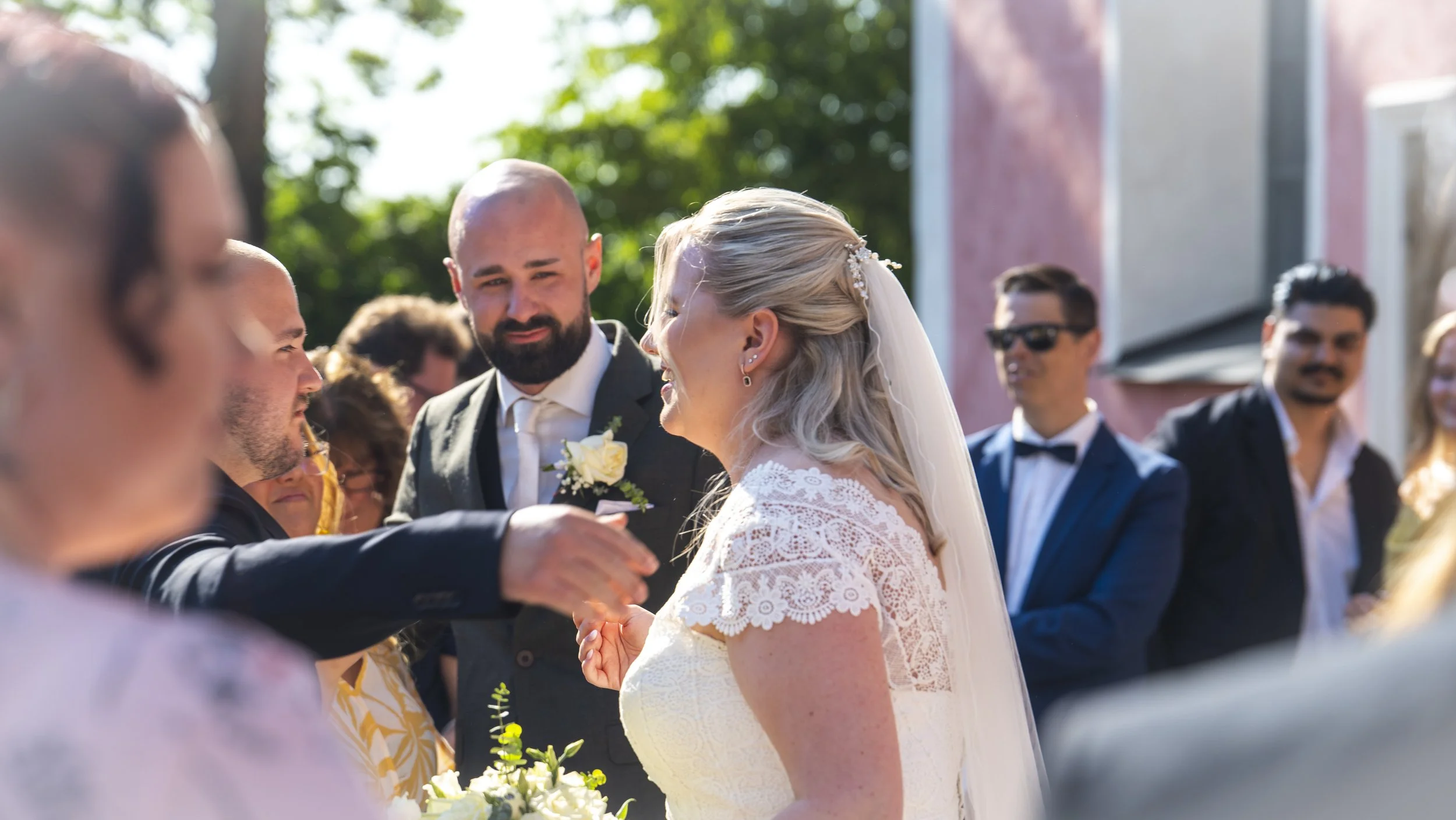 En brud och en man i ett bröllopsceremoni, där andra gäster står omkring utomhus i solen.