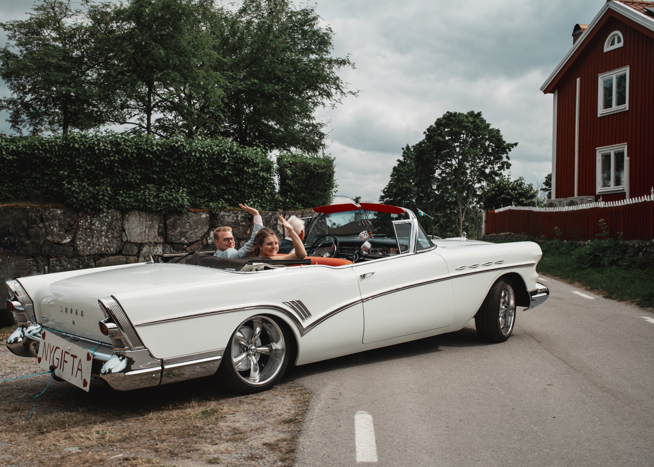 En vit kylig konvertibel med tre personer, två vuxna och en barn, som åker på en landsväg under en molnig dag. Bilen har ett bröllopskännetecken med texten 'NYGIFTA' på baksidan.