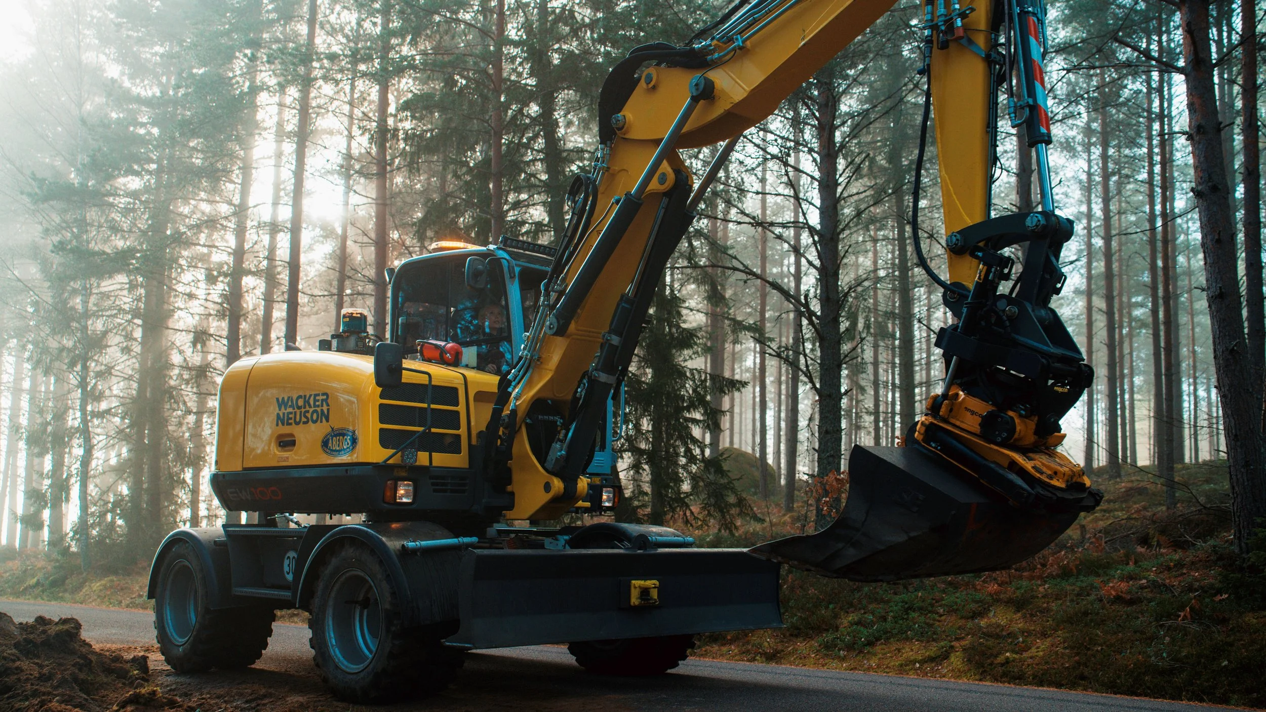 En gul grävmaskin arbetar i en skog under dimmiga förhållanden.