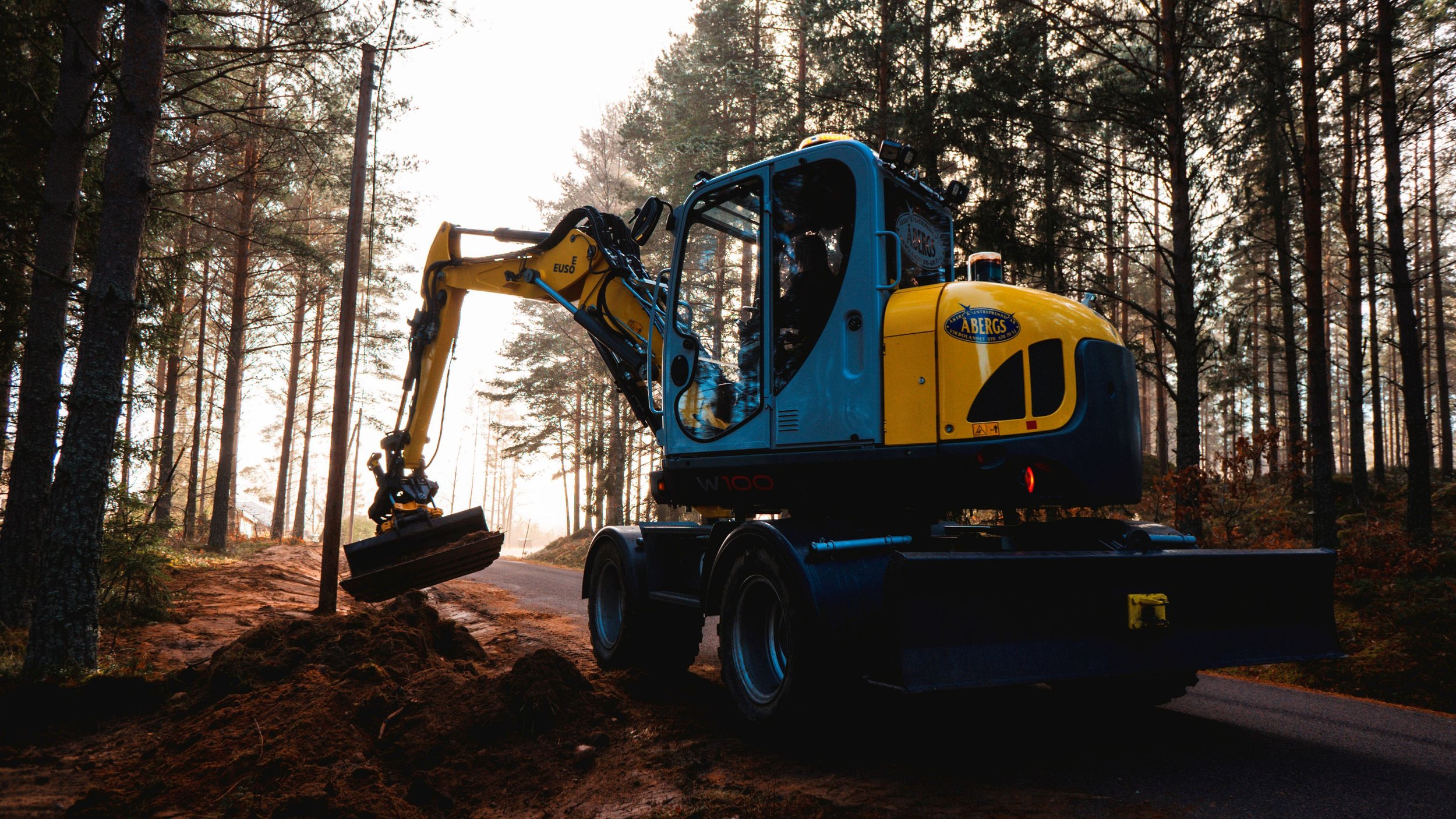 En liten grävmaskin arbetar vid en skogsstig, grävarbete i ett skogsmiljö med träd och jordbank, tidig morgon eller sen eftermiddag.