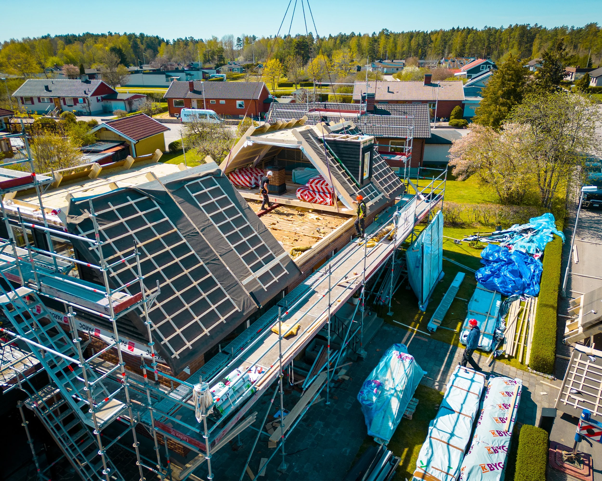 Byggarbetare arbetar på ett hus under konstruktion, med byggnadsställningar och takstolar, i en bostadsområde på vårkanten.