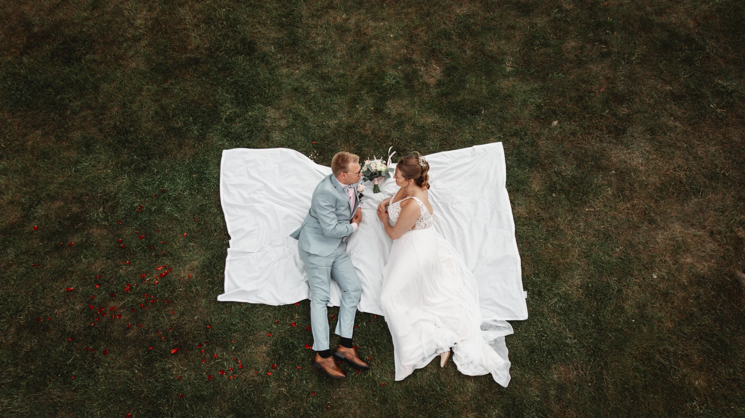 En nygift brud och brudgum ligger på en vit linneduk på gräset, tittar på varandra, med en bröllopsbukett mellan dem.