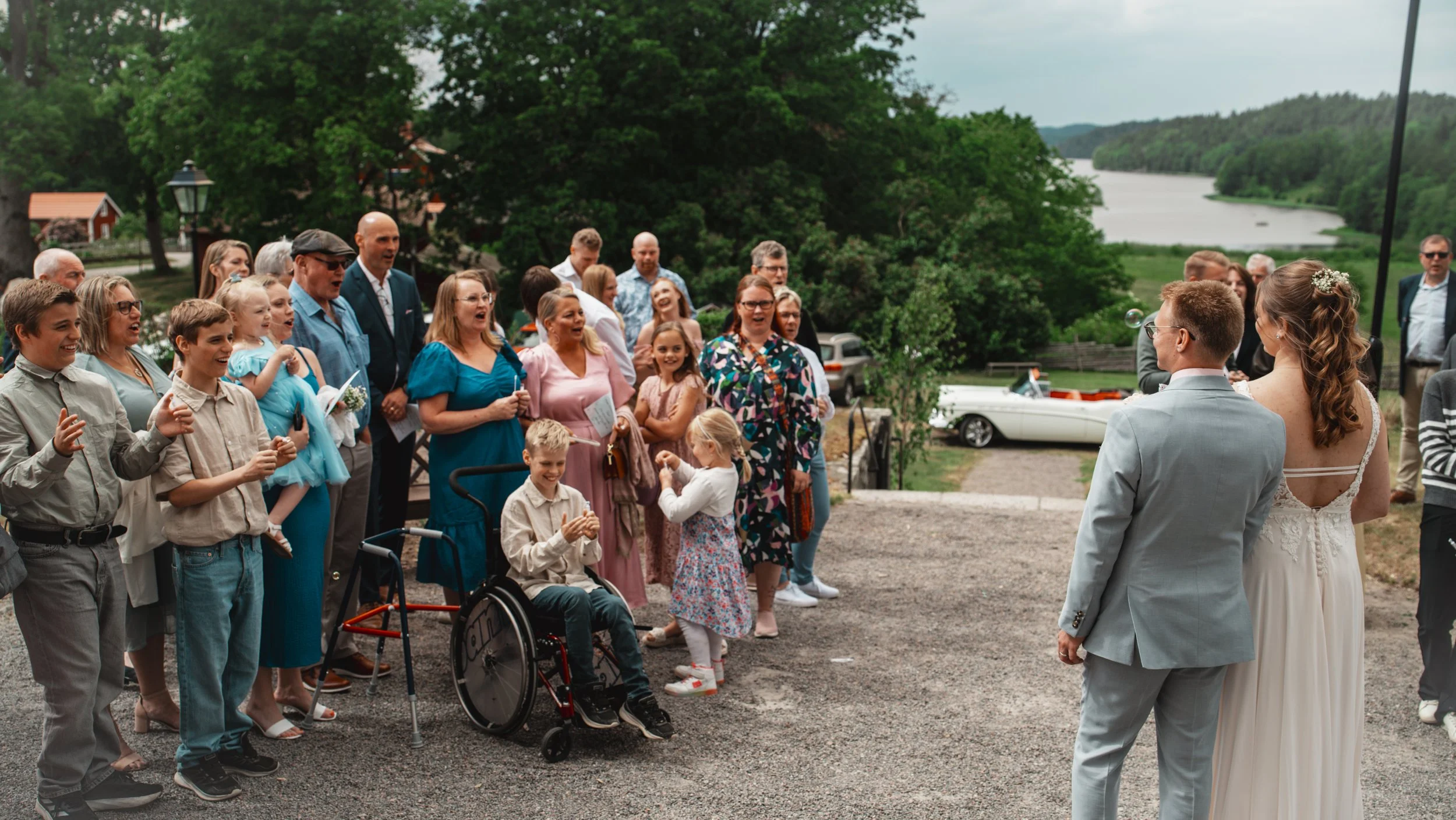 En grupp människor, inklusive barn och vuxna, samlas utomhus för ett bröllop, med en vacker utsikt över en sjö och gröna träd i bakgrunden.