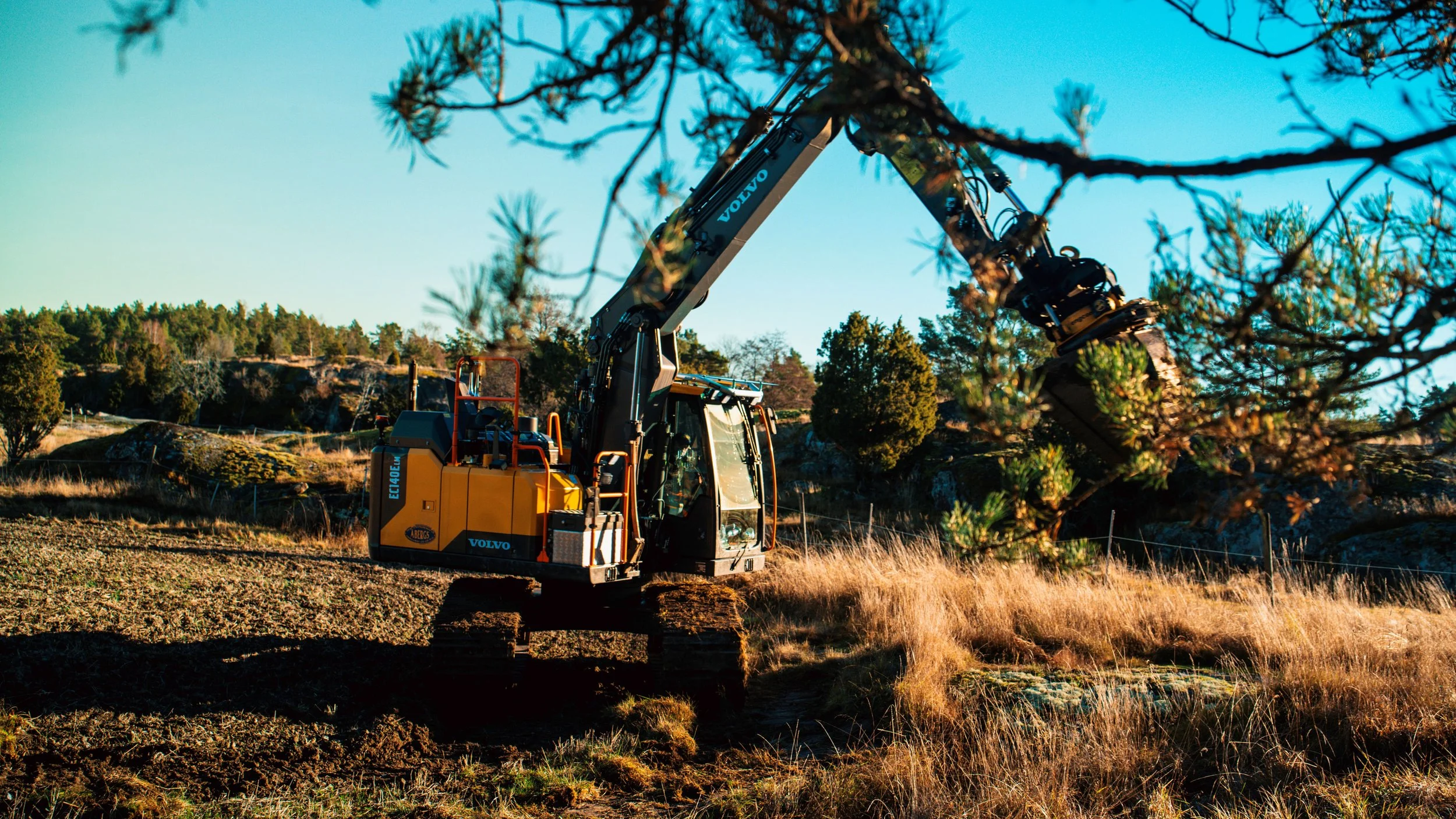 En gul väganläggningsmaskin märkt Volvo och Engcon arbetar i ett terrängområde med träd och gräs.