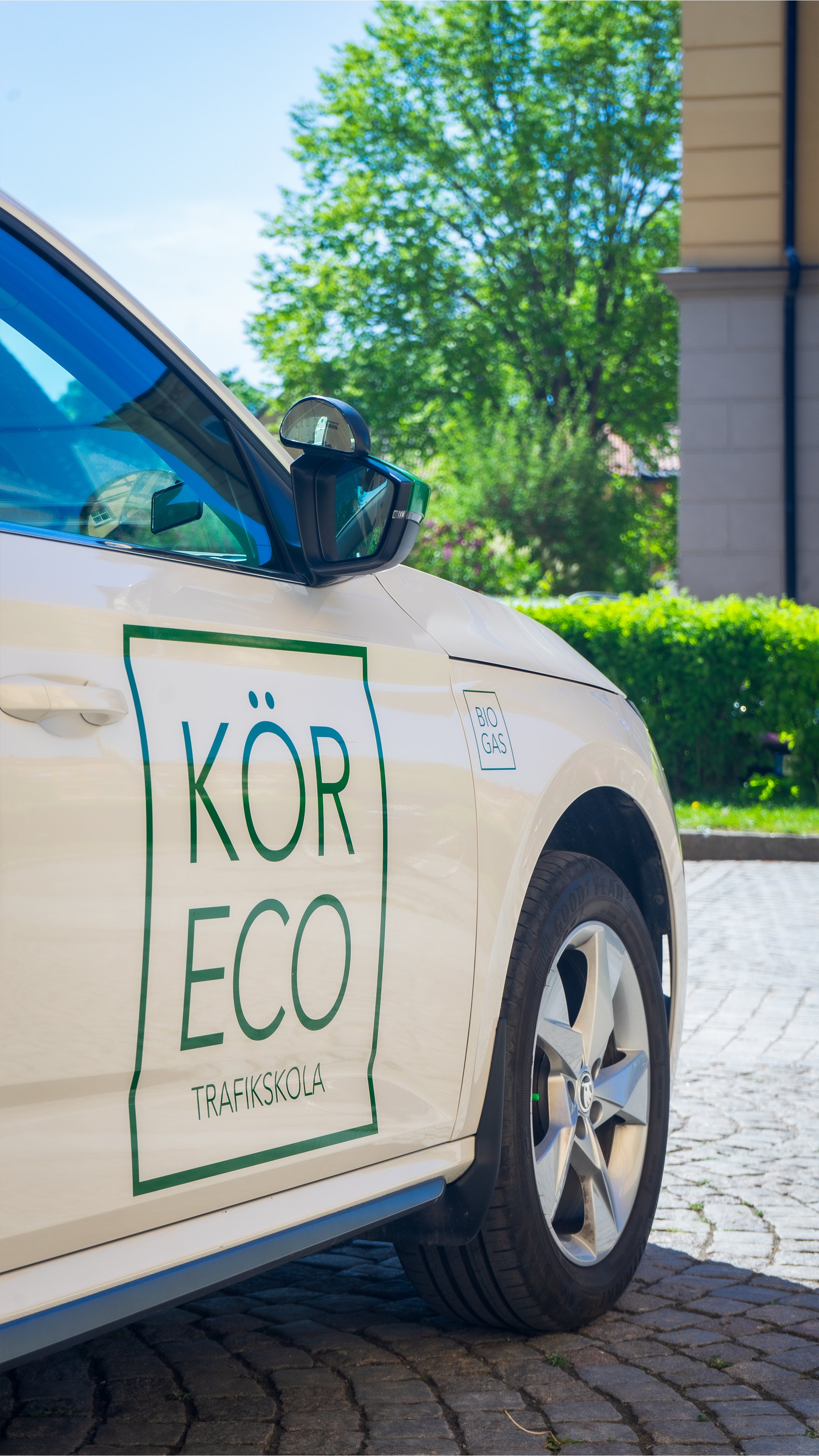 Ett vitbil med etiketten 'Kör ECO Traffikskola' parkerad på en gatukant med grön träd och ett grått hus i bakgrunden, soligt väder.