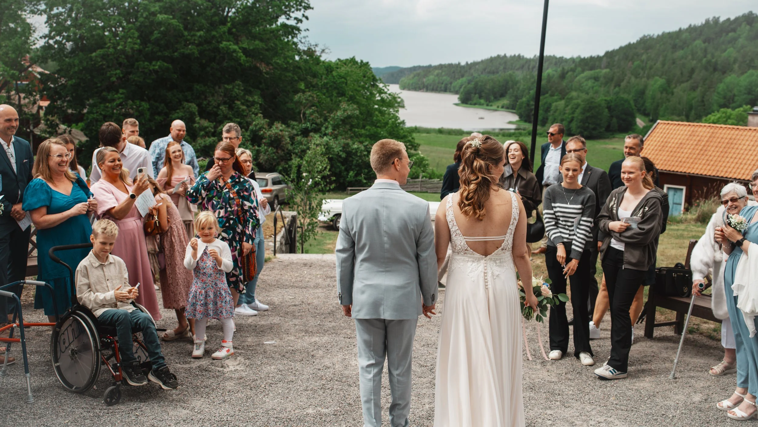 Brudpar framför gäster vid ett utomhusbröllop med grönskande landskap och vatten i bakgrunden.