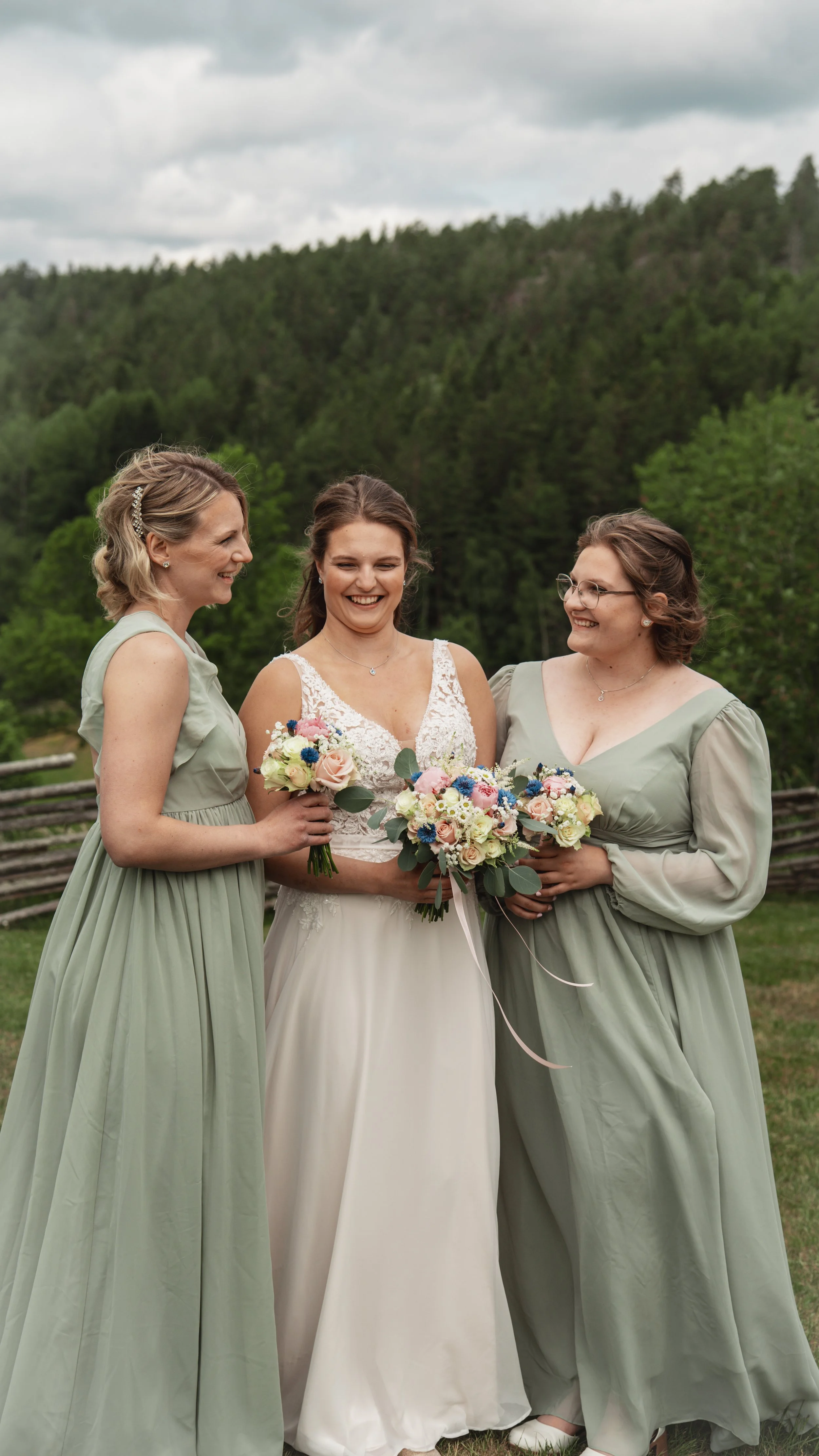 Tre kvinnor på bröllop: en brud i vit klänning och två bridesmaids i ljusgröna klänningar, alla håller buketter av blommor, står utomhus mot en grön skog med molnig himmel.