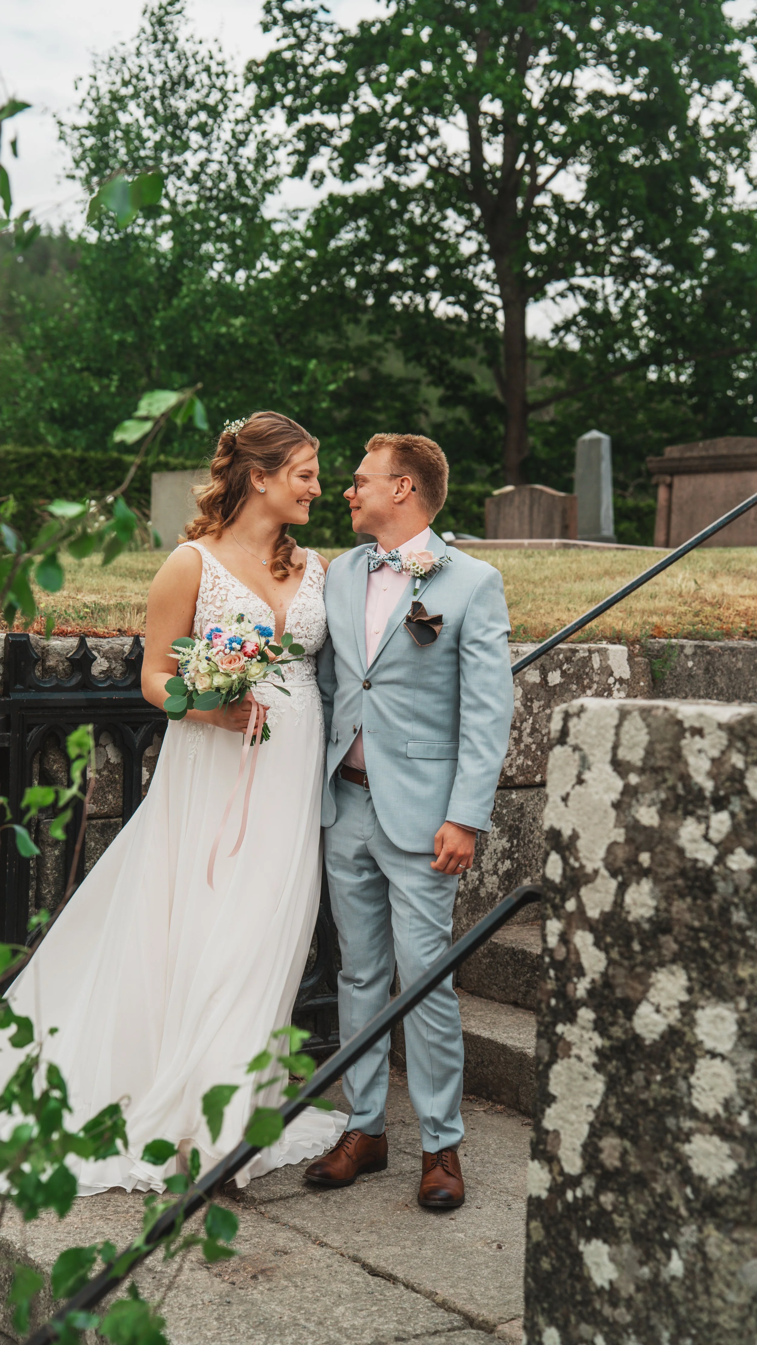 En brud och en brudgum står nära varandra utomhus, ler mot varandra, med en kyrkogård i bakgrunden.