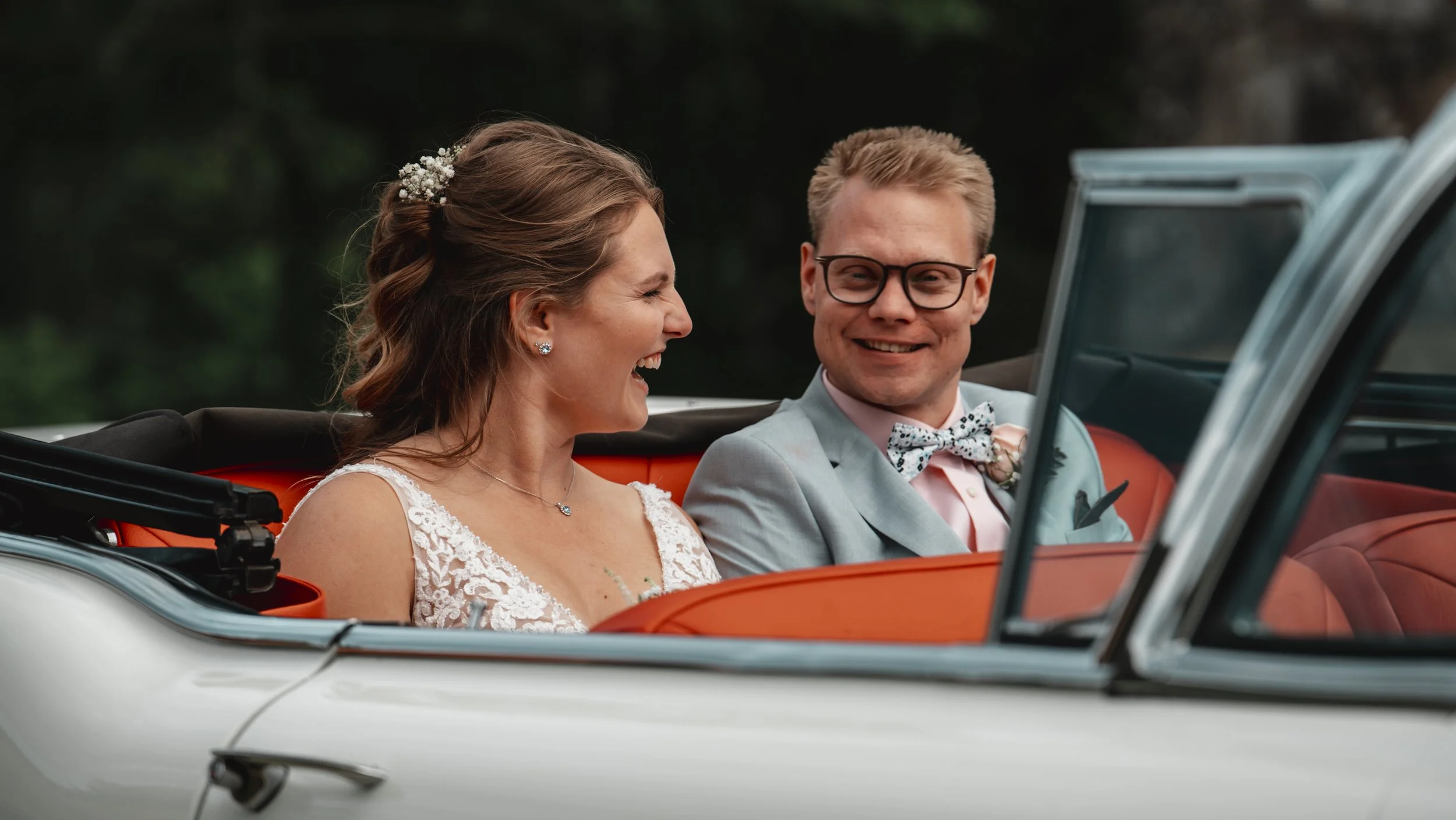 En nygift brud och brudgäst som sitter i en vit vintage konvertibel bil. De ler mot varandra och är klädda i bröllopskläder, bruden i spetsklänning och brudgummen i ljus kostym med fluga. Bakgrund av suddig natur.