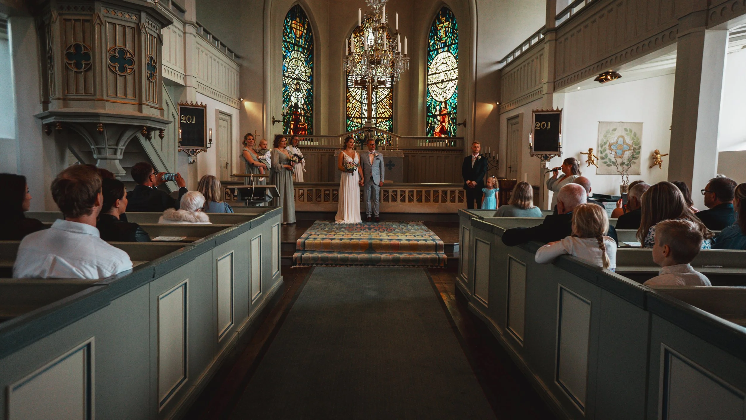 Bröllopsceremoni i ett kyrkorum med målade glasfönster bakom altaret, brud och brudgum står framför prästen, omgiven av brudtärnor och marskalkar, med gäster sittande i bänkar.