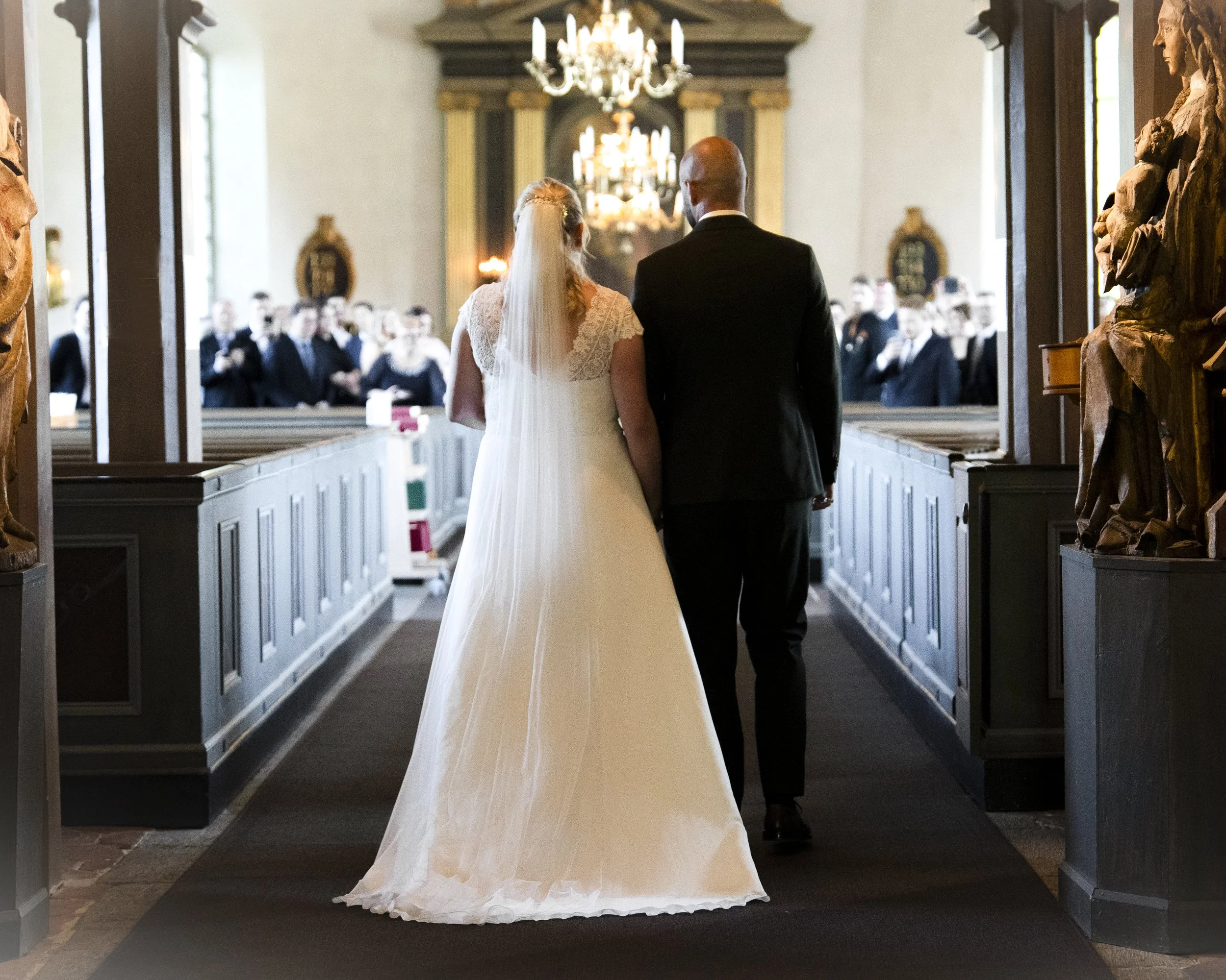 En brud och en brudgum som håller varandra i handen i kyrkan under deras bröllopsceremoni, med gäster som bevittnar i bakgrunden.