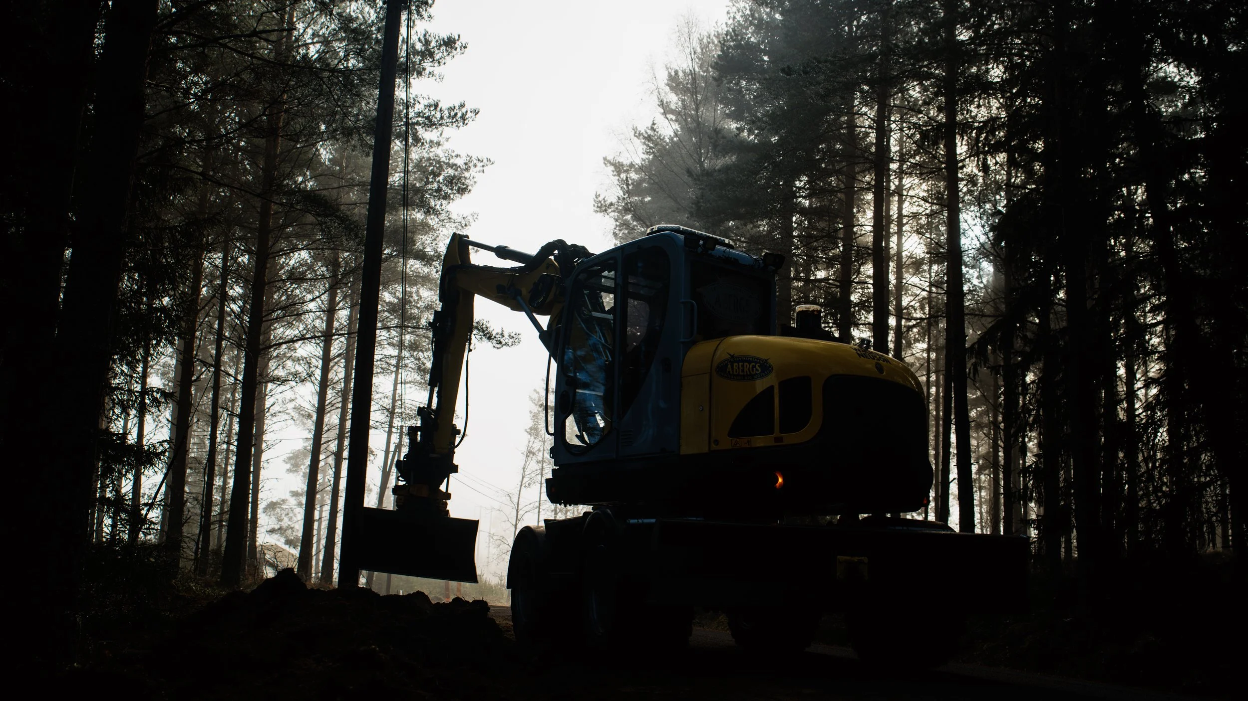 En skuggbild av en grävmaskin i en skog med höga träd, mot en ljus himmel.
