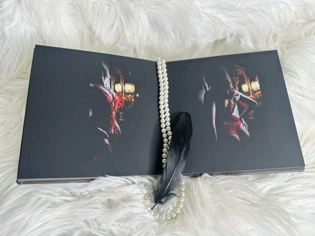 Open book with dark photographs of women wearing red lingerie, adorned with a pearl necklace and a black feather, placed on a white furry surface.