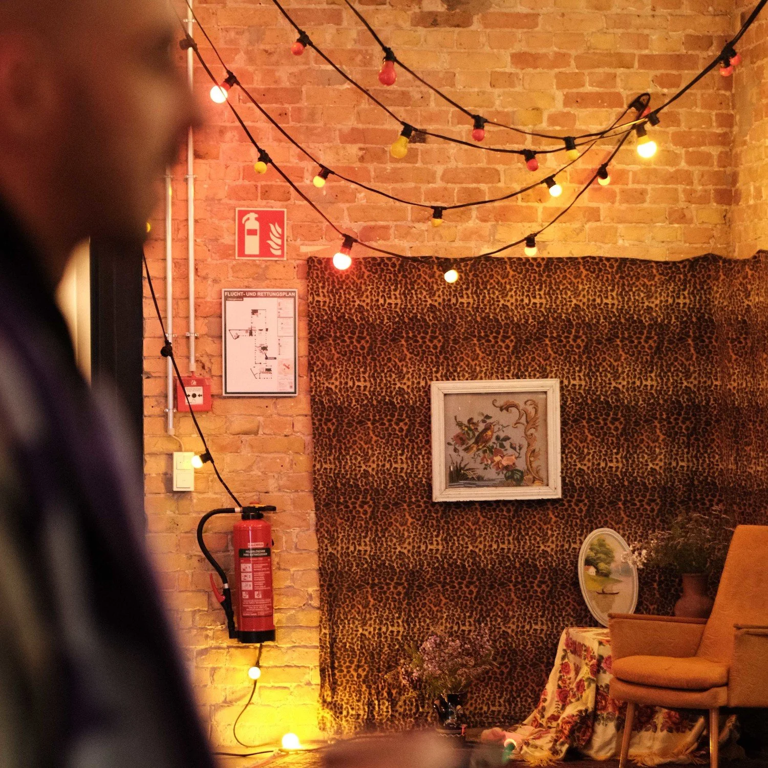 Interior scene with string lights hanging from a brick wall, a framed picture of birds, a table with a floral cloth, a yellow chair, and a leopard print backdrop. A fire extinguisher and fire safety signs are also visible.