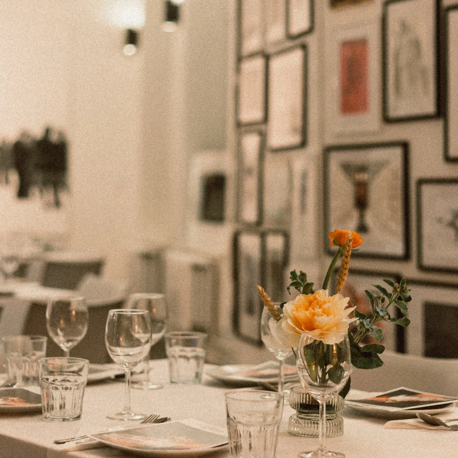 A table set for a meal with multiple glasses and plates, with a flower centerpiece in a glass vase, against a wall decorated with framed pictures.