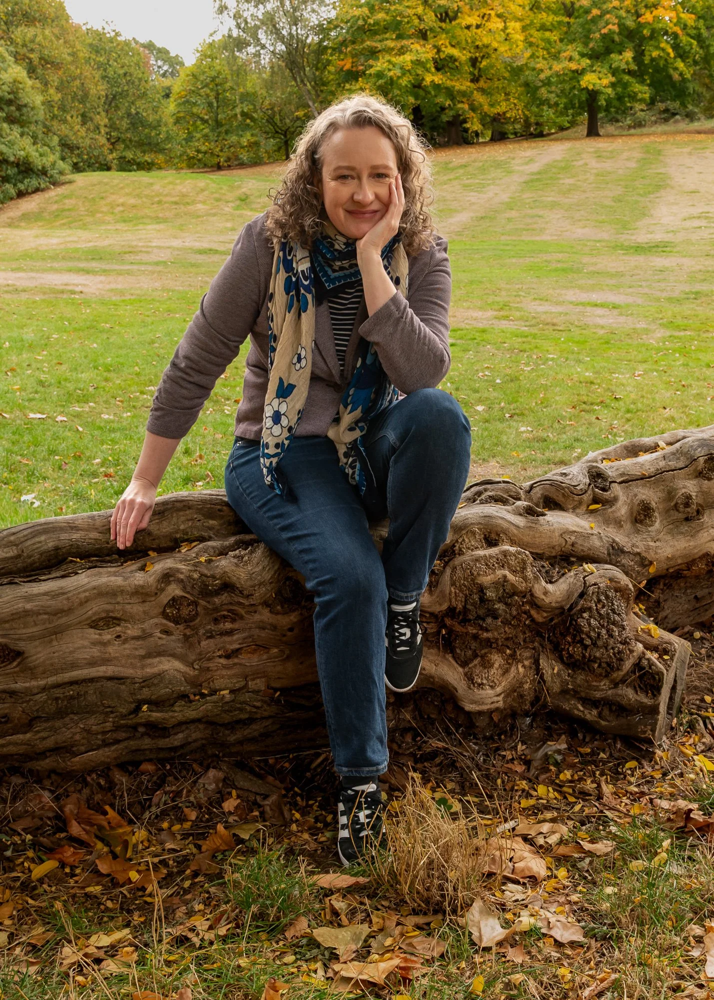 A woman with curly gray hair sitting on a fallen tree trunk in a park with green and yellow trees in the background, smiling with her hand on her cheek.