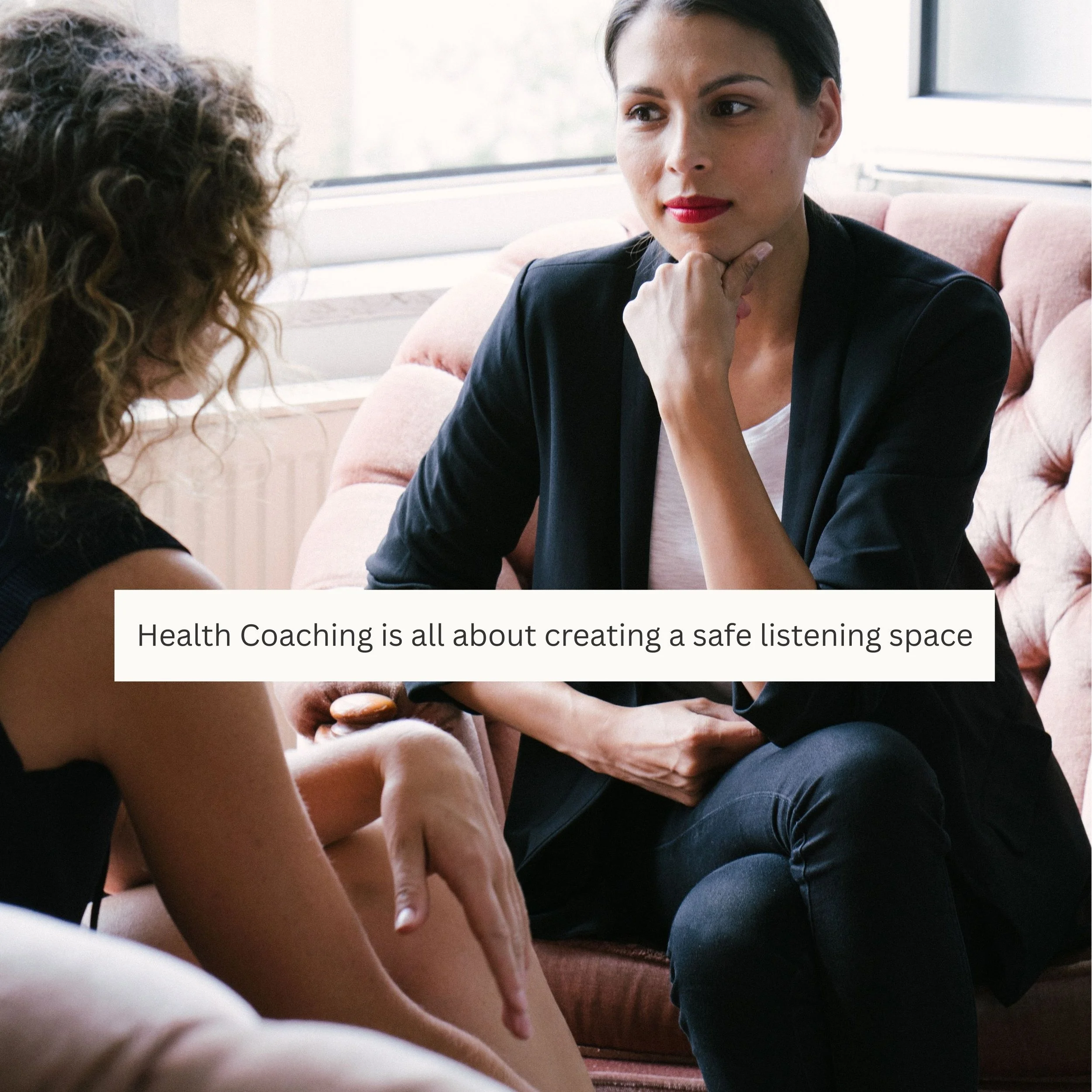 Two women having a conversation in a cozy, well-lit room with a pink sofa. The woman on the right is listening attentively, resting her chin on her hand. The woman on the left is speaking, and a caption reads: "Health Coaching is all about creating a safe listening space."