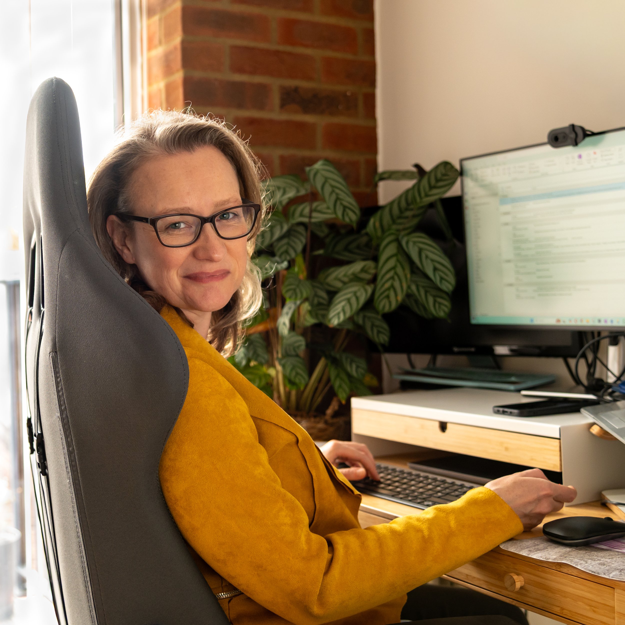A woman with glasses sitting at a desk in front of a computer, wearing a mustard yellow blazer, in a room with a brick wall, window, and large potted plant.