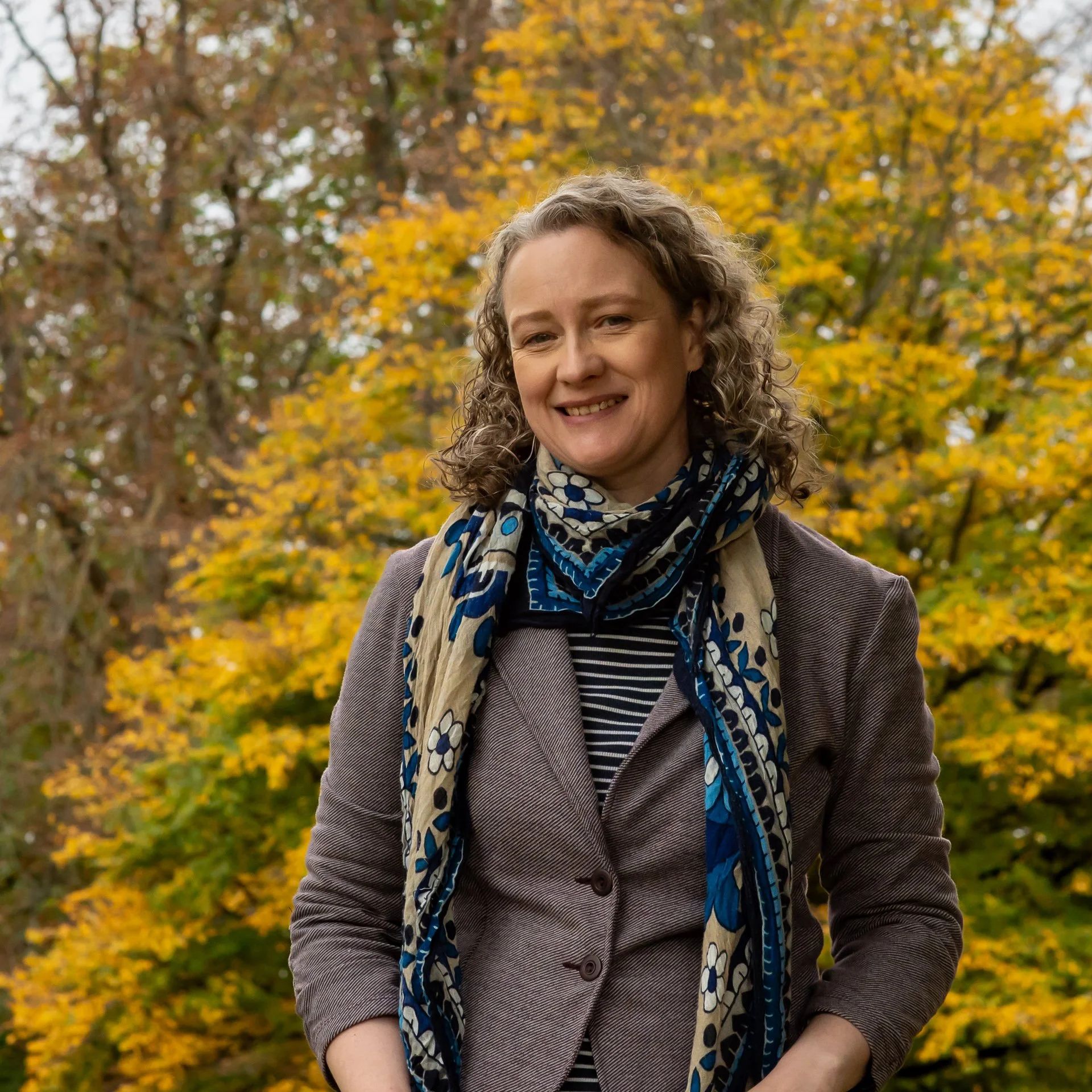 A woman with curly hair standing outdoors during fall, smiling, wearing a brown blazer, striped shirt, and a blue patterned scarf with autumn trees with yellow leaves in the background.